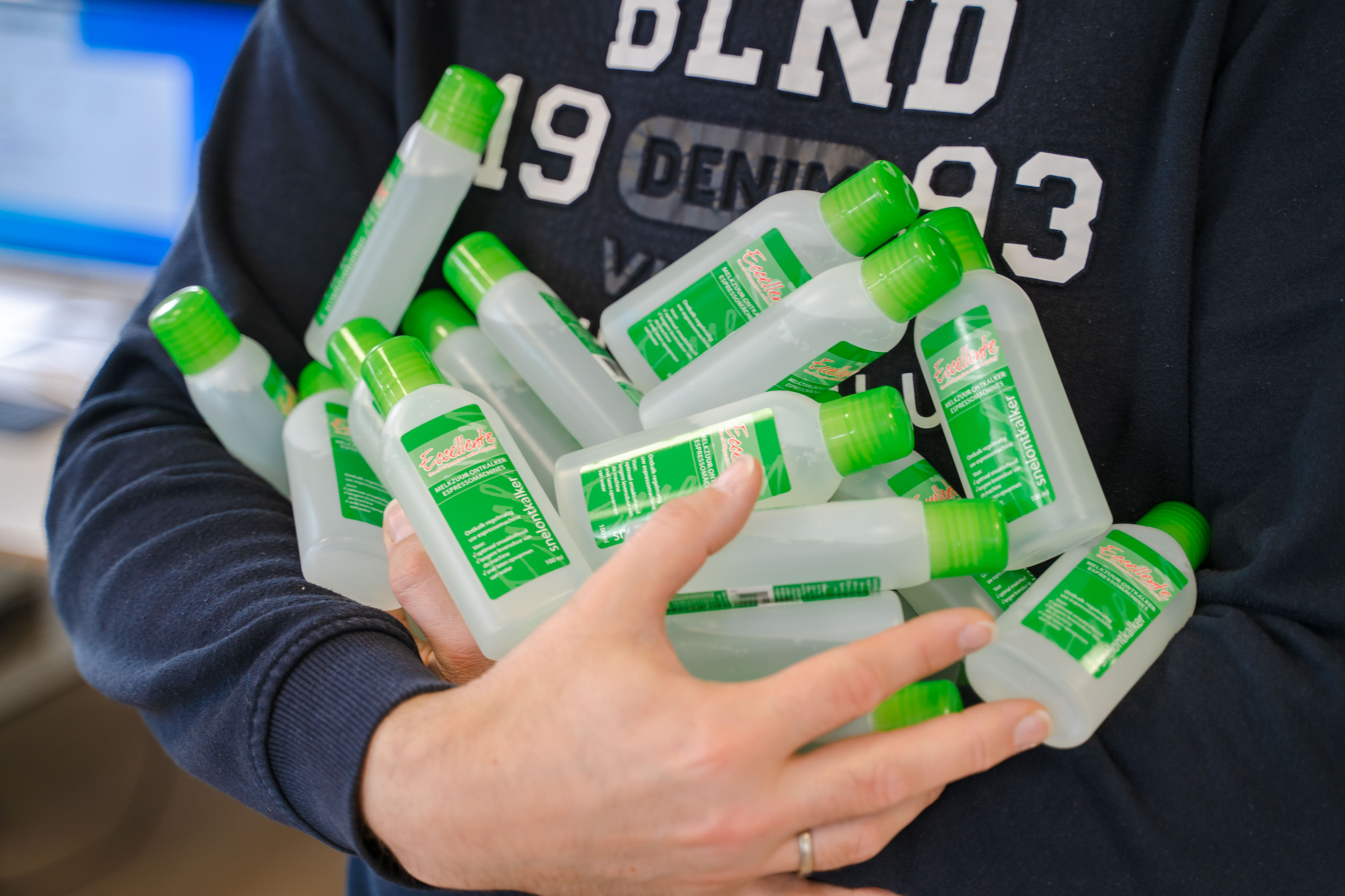 A person holds multiple white bottles with green caps. Bottles have labels with green text. Background is blurred.