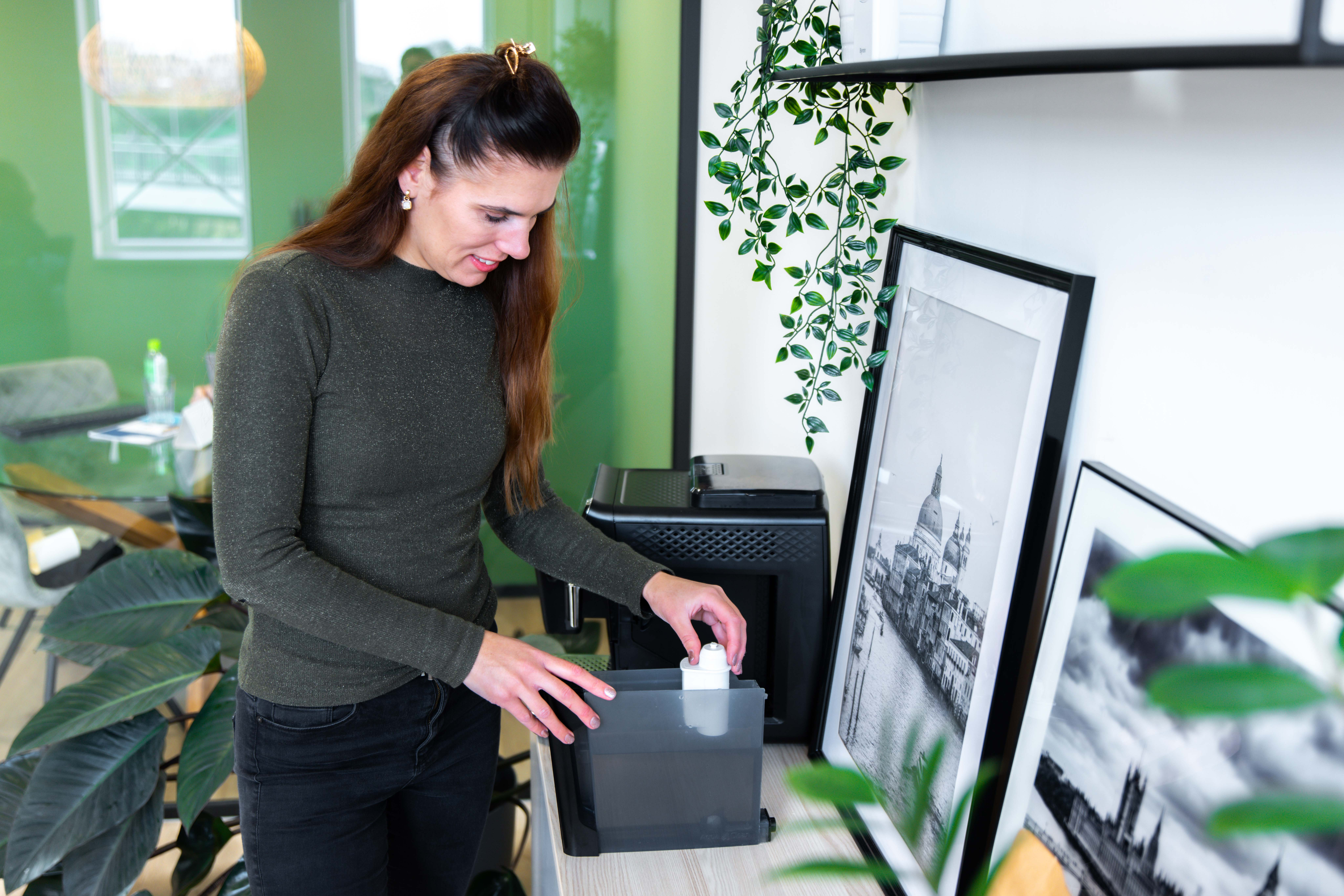 Een vrouw in een groene top vult een koffiezetapparaat. Een ingelijste foto en planten staan op de achtergrond.