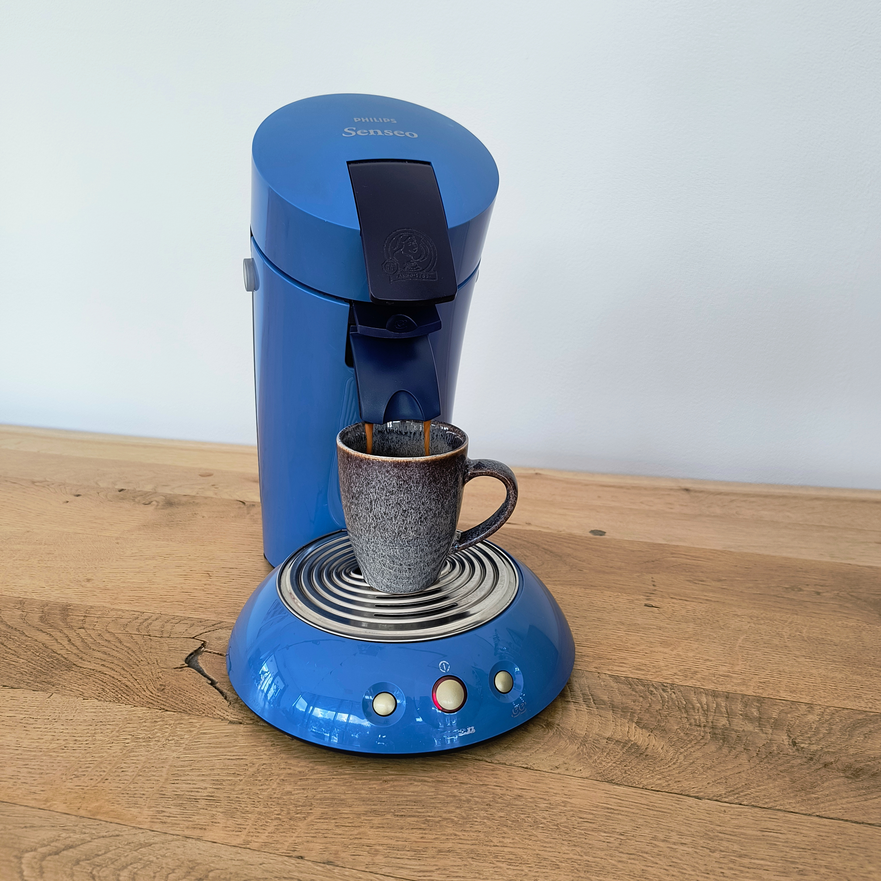 Een blauwe koffiemachine met een kopje, koffie aan het zetten. Het staat op een houten oppervlak, met een witte muur erachter.