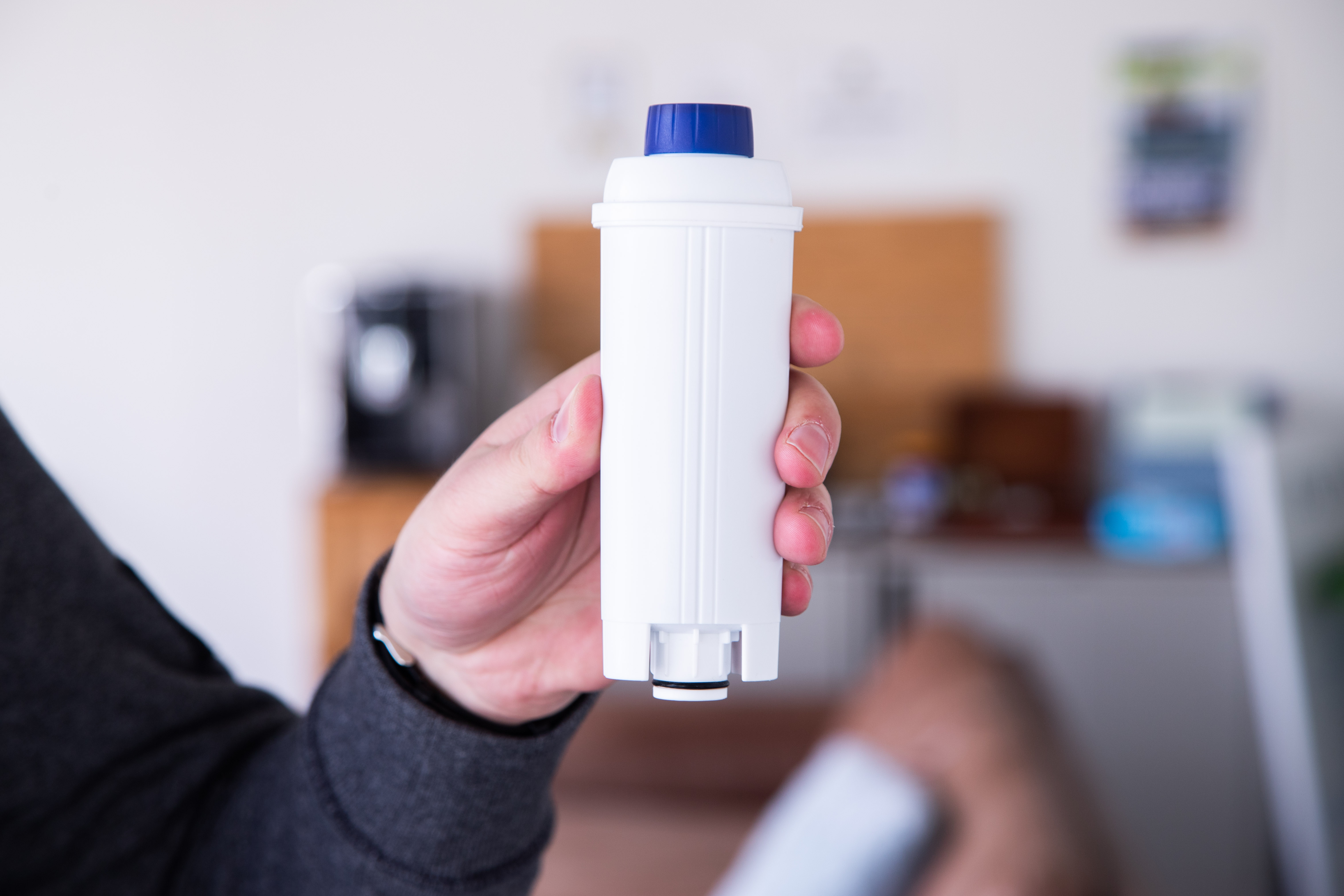 Hand holding a white water filter with a blue cap. Blurred background includes furniture.