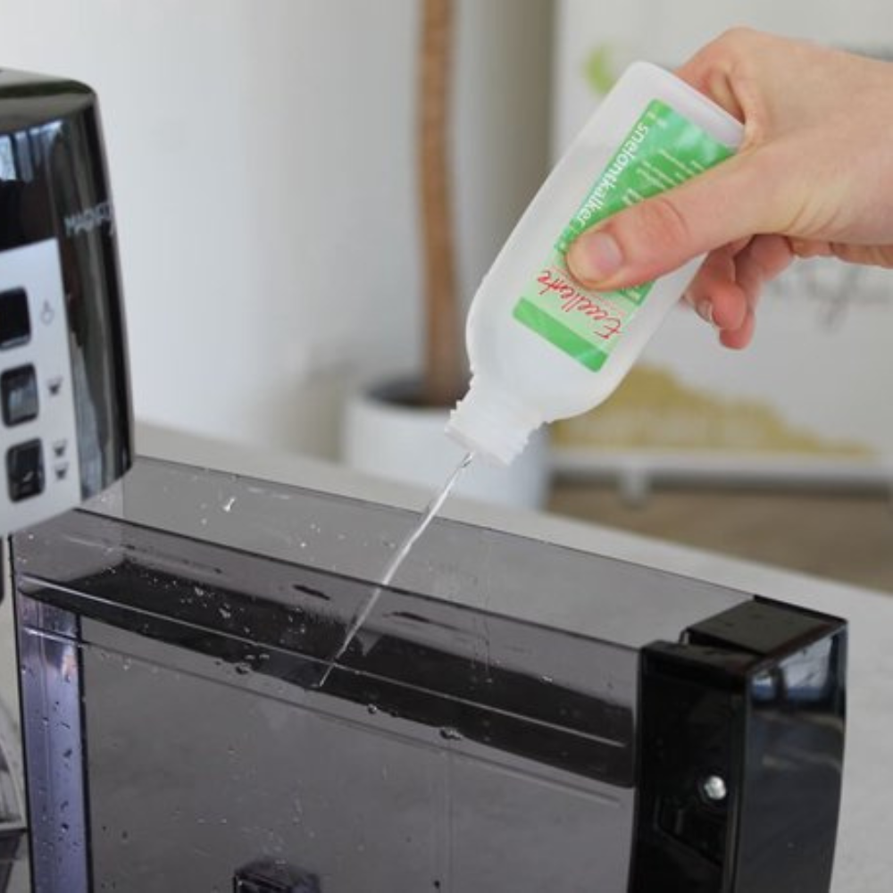 Une main verse du liquide d'une bouteille blanche étiquetée 'Excellente' dans un réservoir d'eau noir d'une machine à café.
