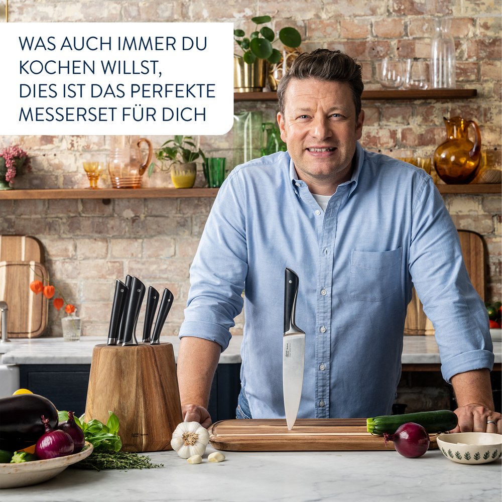 Koch Jamie Oliver mit einem Messerset. Verschiedenes Gemüse und Knoblauch liegen auf der Theke.