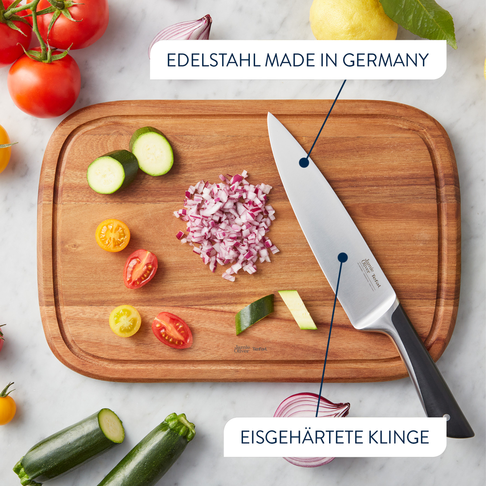 Ein Schneidebrett mit Messer, gehackten roten Zwiebeln, Zucchinischeiben und Tomaten. Das Messer hat einen schwarzen Griff.
