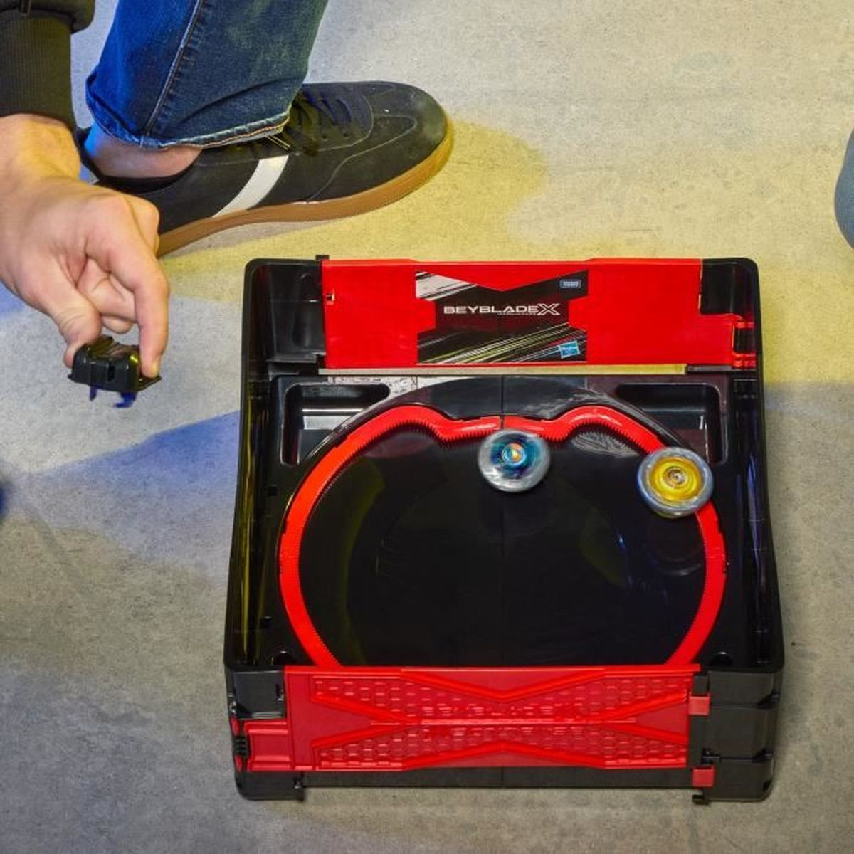 Una persona sostiene un lanzador Beyblade negro sobre una arena Beyblade. Dos Beyblades giran dentro de la arena negra con borde rojo.