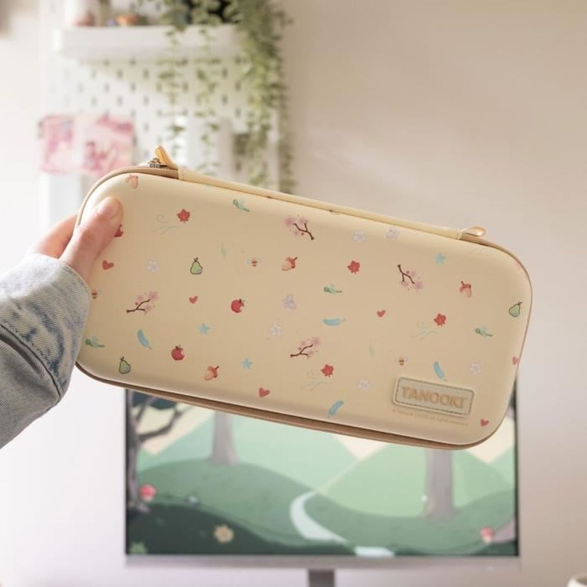 Una mano sostiene una funda beige con diseños florales. La funda dice 'Tanooki'. El fondo incluye decoración y una pantalla.