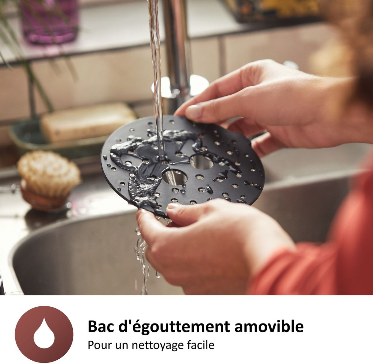 Ręce myją czarną, perforowaną część urządzenia kuchennego pod kranem. Woda spływa w dół, z zlewem i innymi elementami w tle.