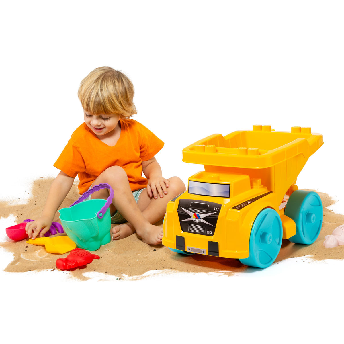 Un niño juega con juguetes de arena, incluyendo un camión volquete amarillo y un cubo verde. El niño lleva una camiseta naranja y está sentado en la arena.