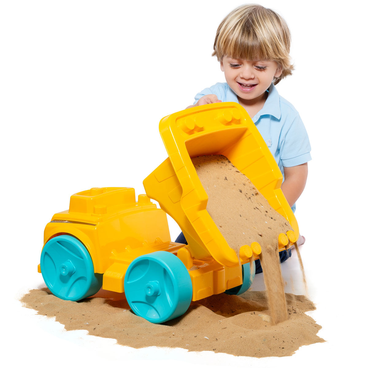 Un niño sonriente vierte arena de un camión de juguete amarillo. El camión tiene ruedas azules. La arena se derrama sobre una superficie blanca.