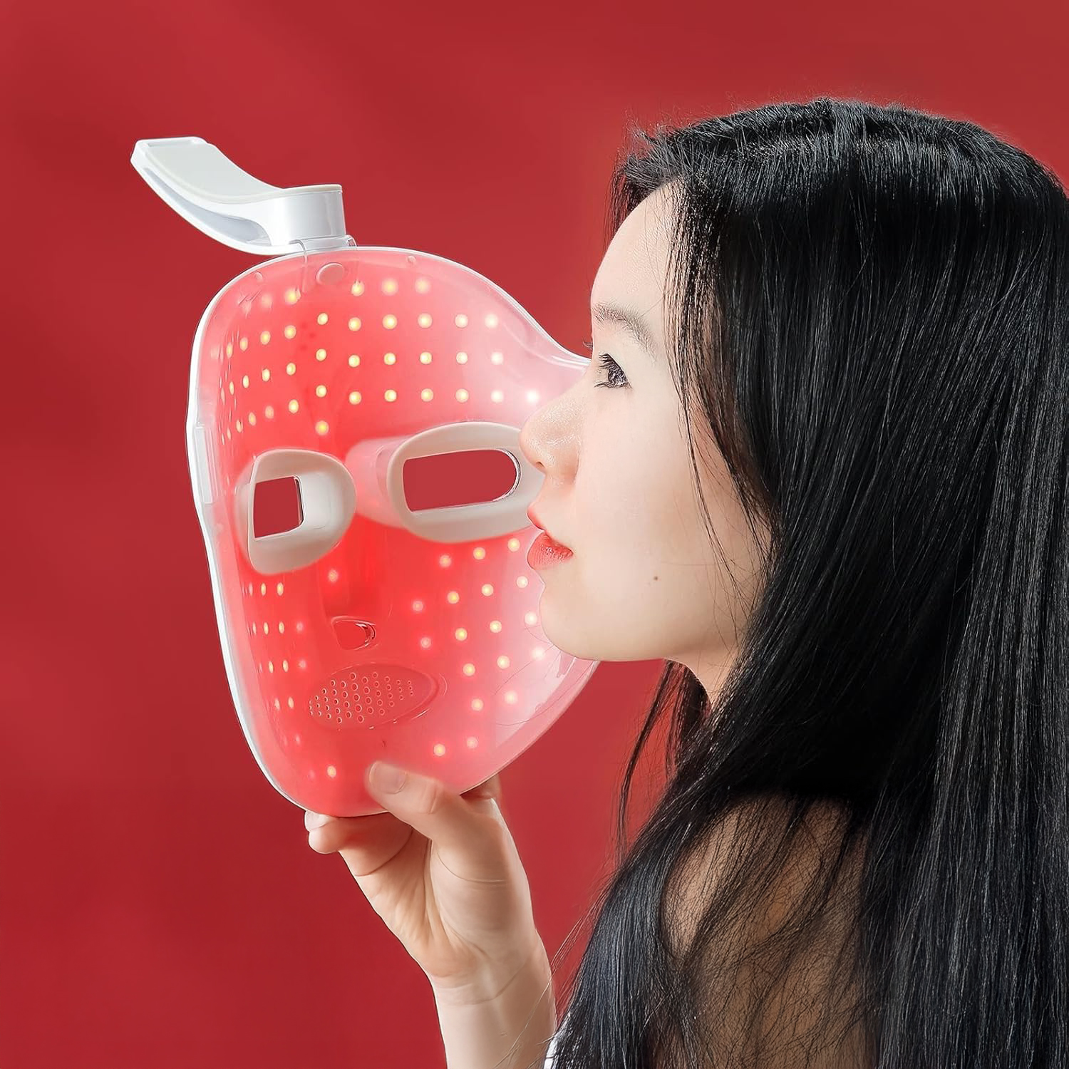 Una donna tiene in mano una maschera facciale a LED rossa. Guarda la maschera, con capelli neri e sfondo rosso.