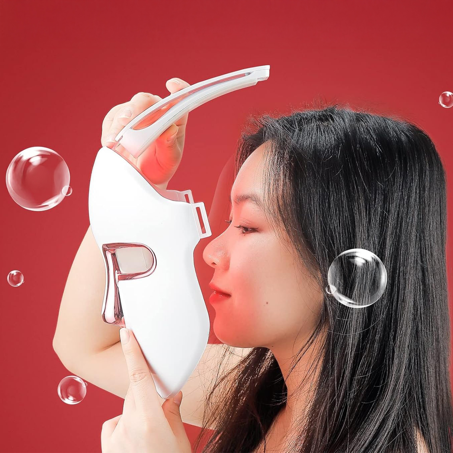 Una donna tiene in mano una maschera facciale a LED bianca. È contro uno sfondo rosso. Bolle fluttuano intorno a lei e alla maschera.