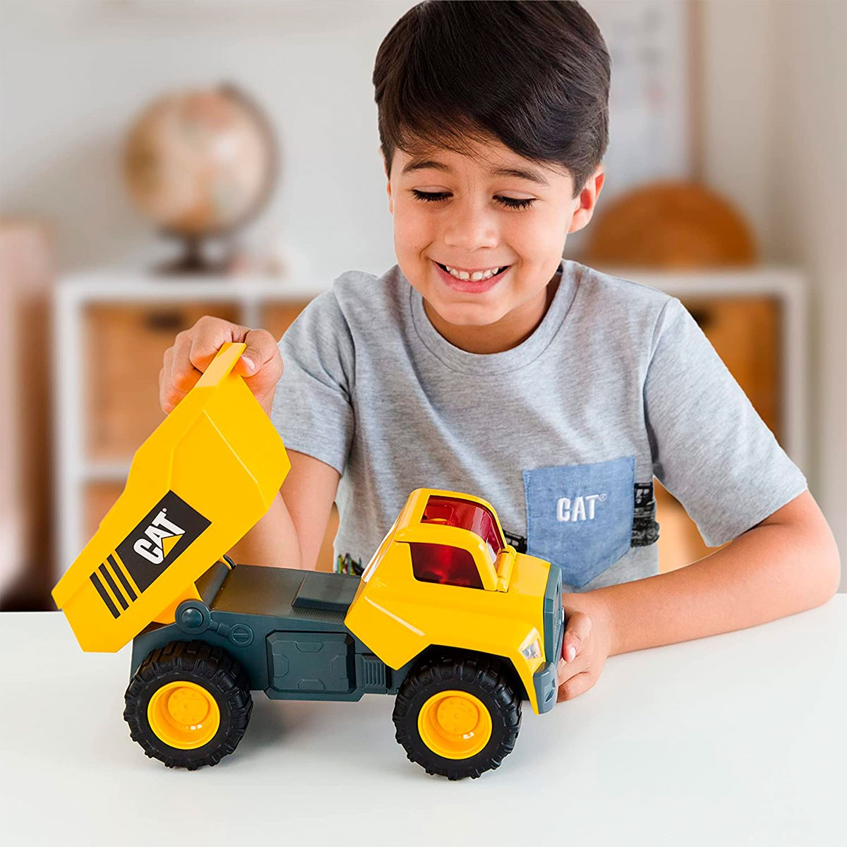 Un ragazzo sorride mentre gioca con un camion ribaltabile CAT giallo e nero. Sta sollevando il cassone del camion. Lo sfondo ha un mappamondo.