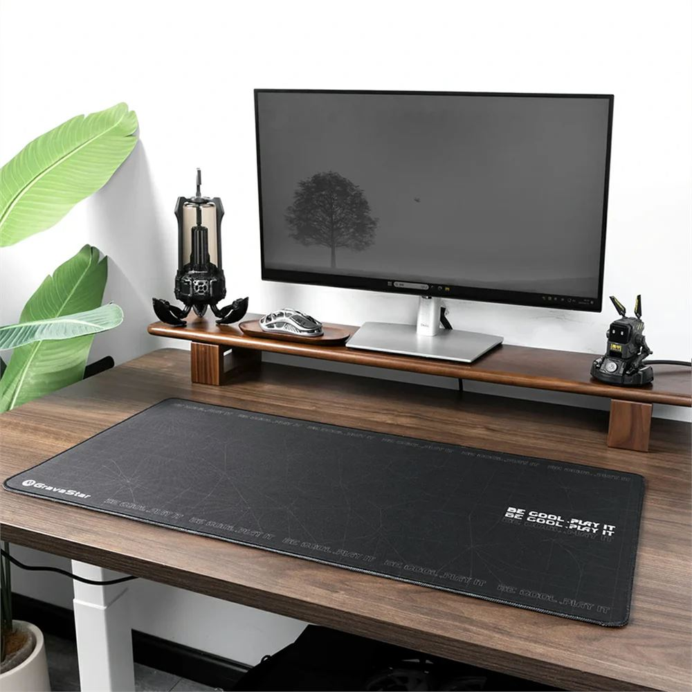 Bir monitör, fare ve siyah bir mousepad ile bir masa. Masada bir bitki ve bazı süslemeler var.