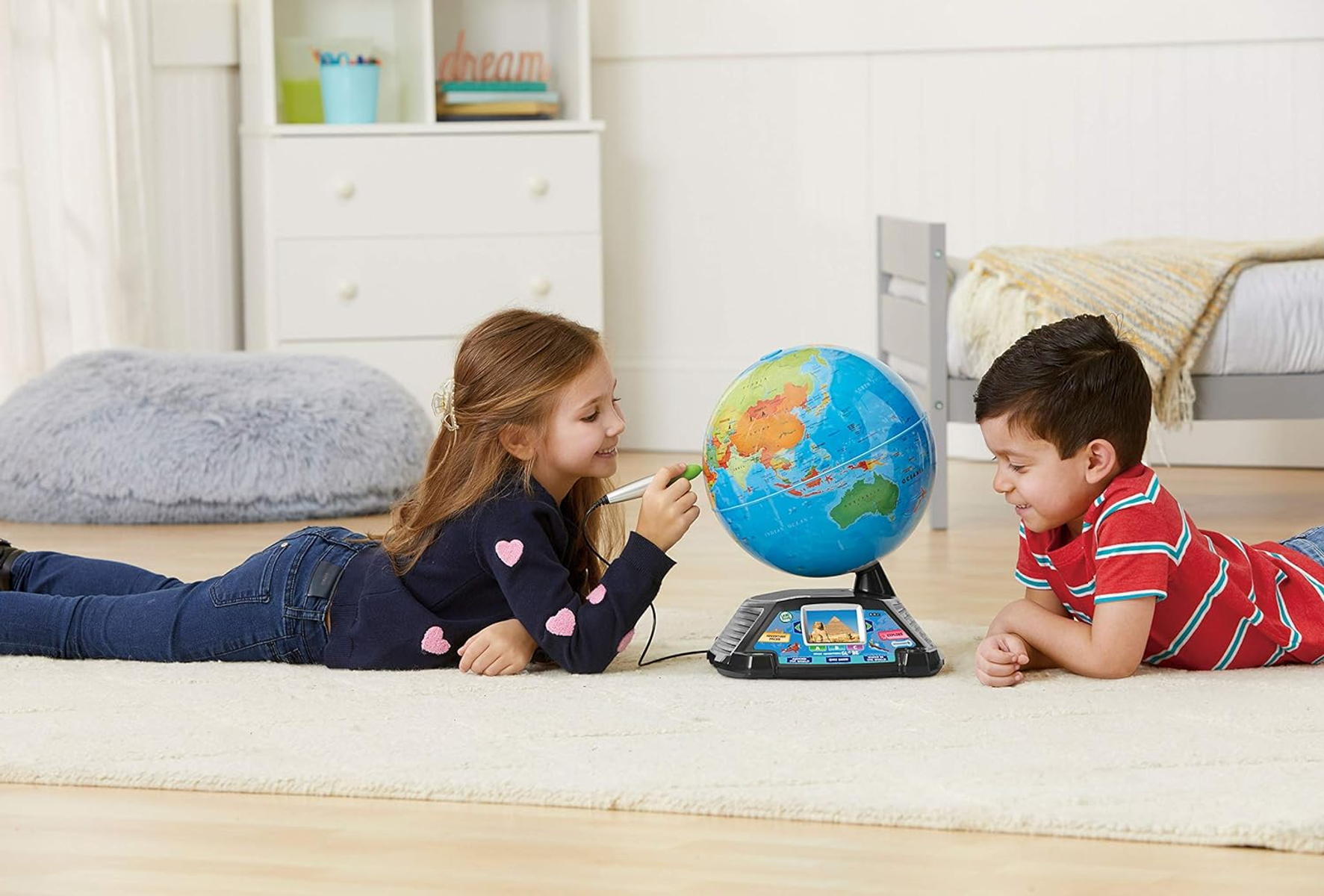 Dos niños estudian un globo terráqueo sobre una alfombra. La niña usa un bolígrafo. El niño mira. Hay una cama y muebles en el fondo.