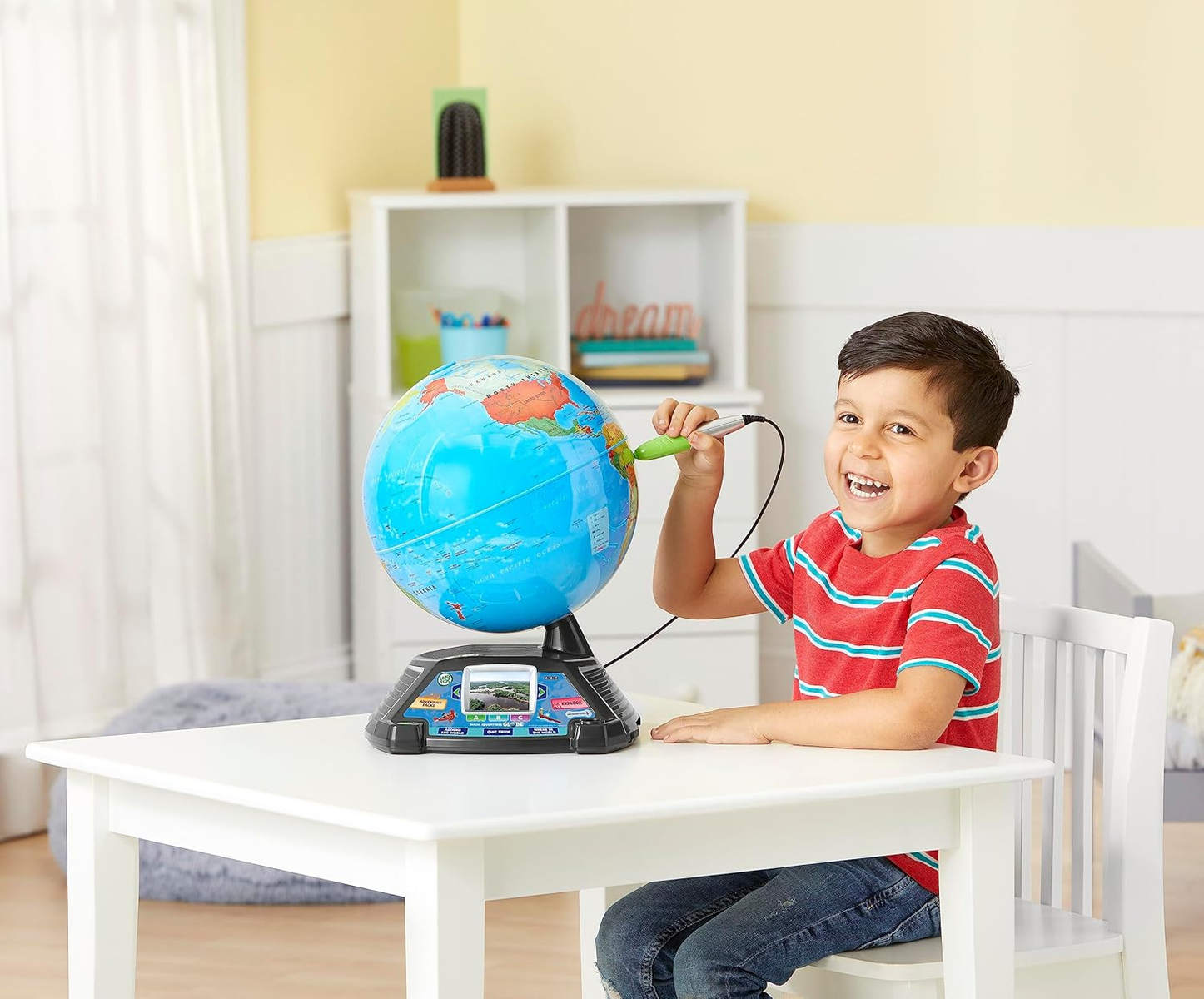 Un niño con una camisa roja usa un bolígrafo en un globo terráqueo. Está sentado en una mesa blanca. Hay una estantería blanca en el fondo.