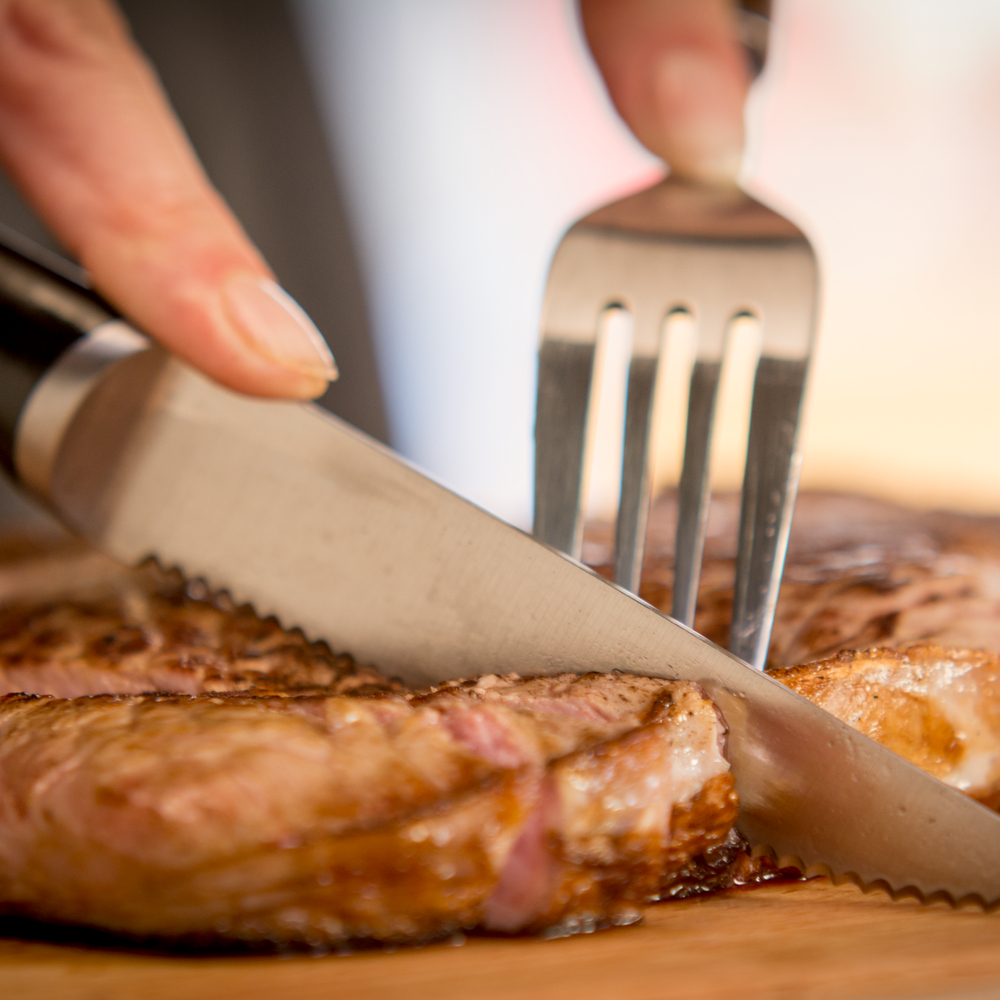 Ręka kroi ugotowany stek nożem z ząbkami, podczas gdy widelec go trzyma. Stek leży na drewnianej desce.