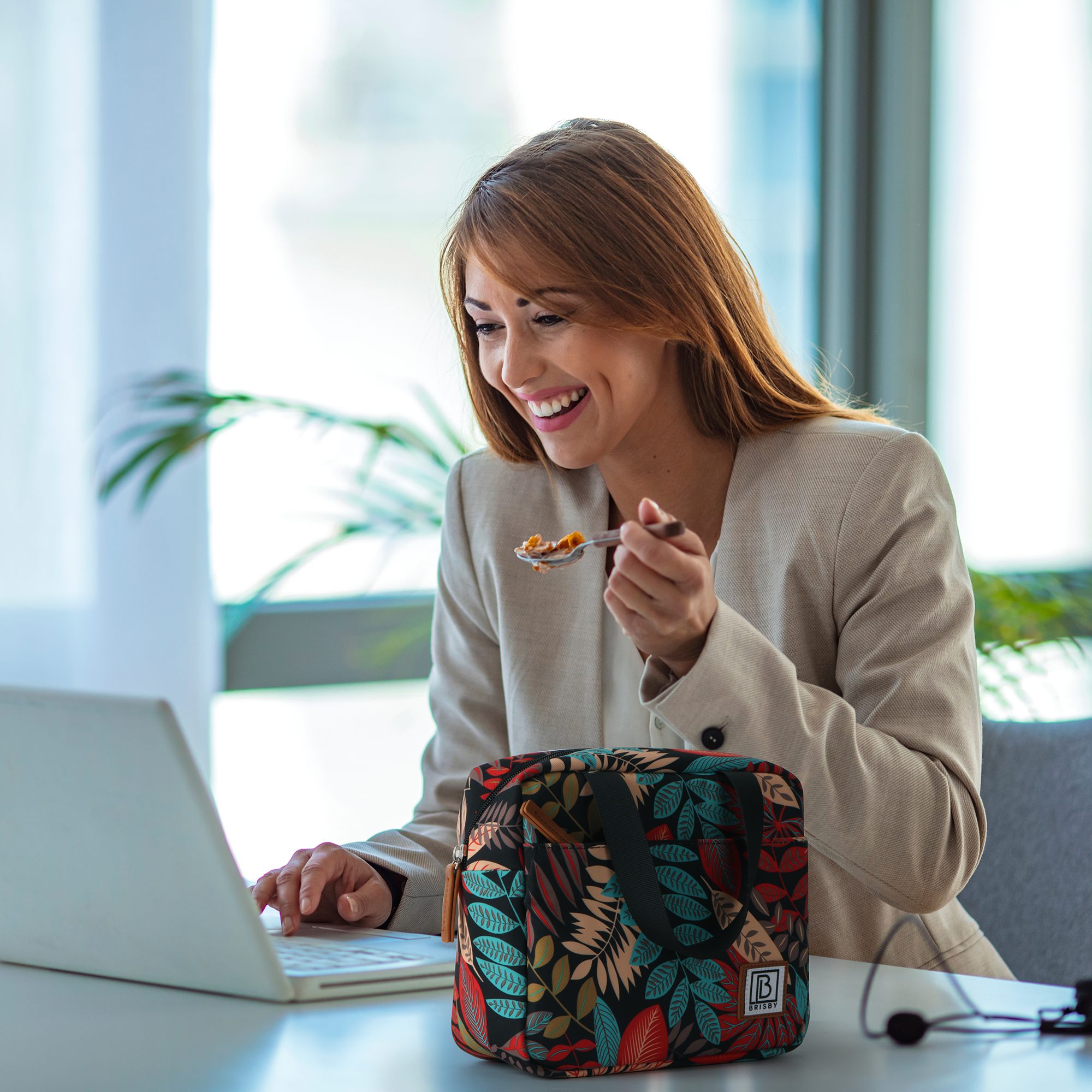 Vrouw in beige blazer eet van een lepel terwijl ze een laptop gebruikt. Een bloemen lunchtas staat op het bureau.