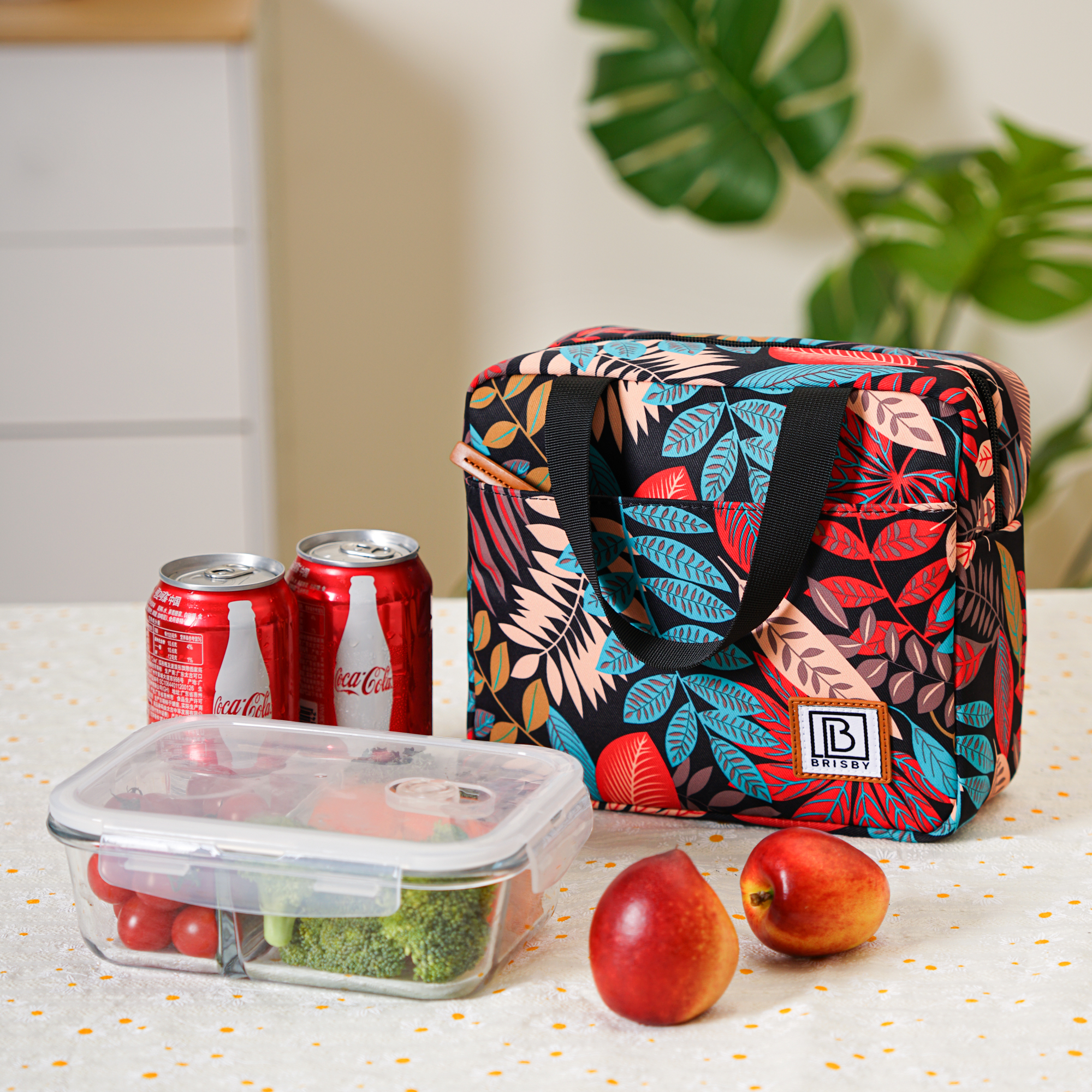 Een lunch met een bloemrijke lunchtas, twee Coca-Cola-blikjes, fruit en een glazen bak met groenten.