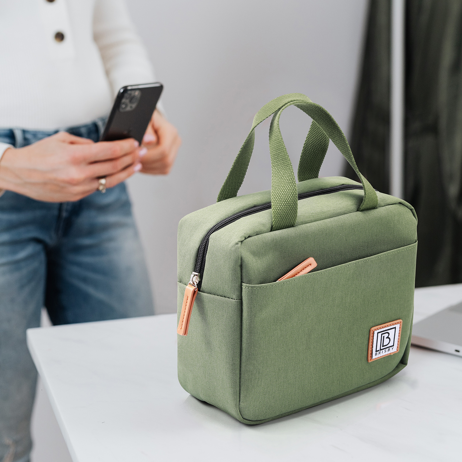 Een groene lunchtas met een leren ritstrekker staat op een witte tafel. Een persoon houdt een telefoon op de achtergrond vast.