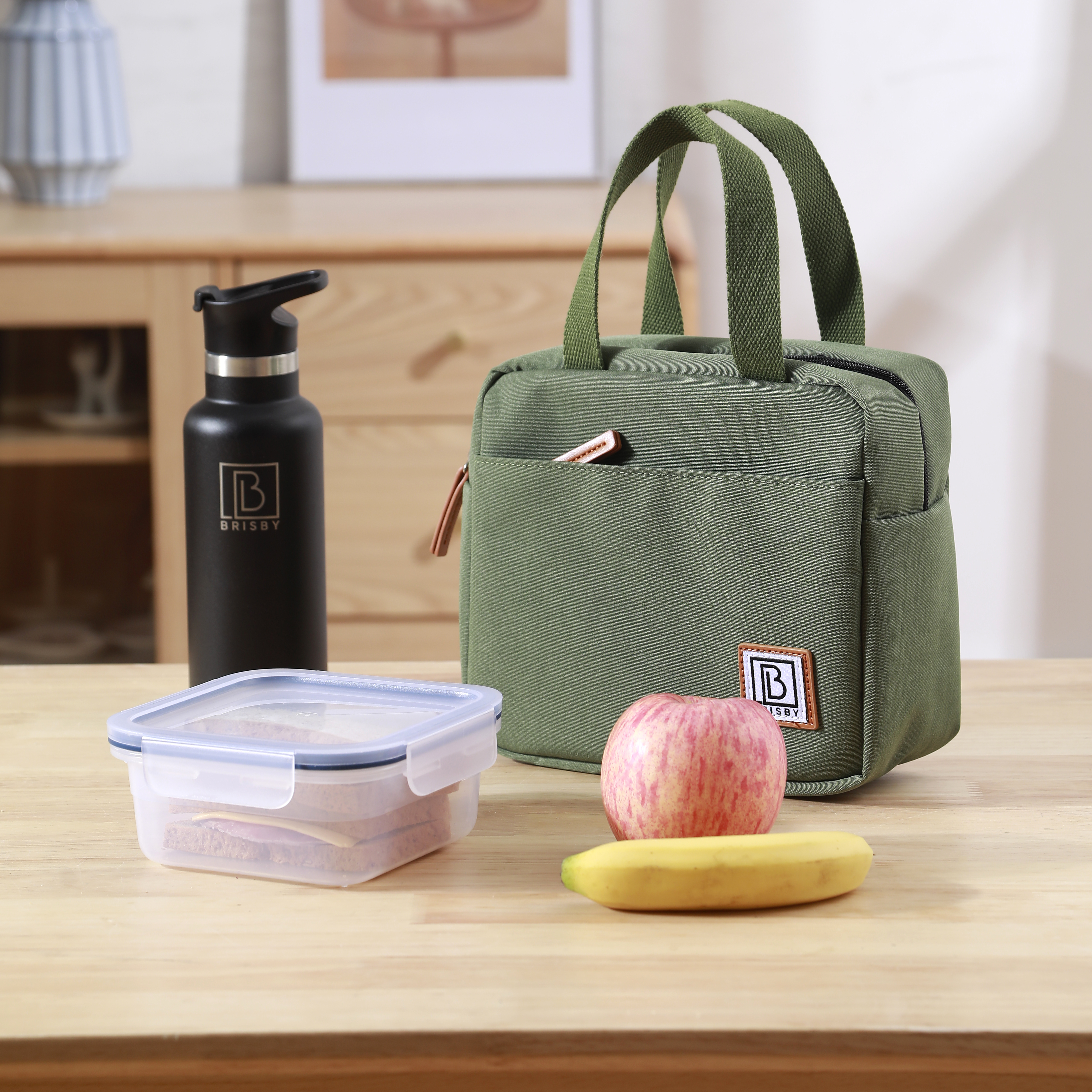Een groene lunchtas, zwarte waterfles, doorzichtige container, appel en banaan staan op een houten tafel.