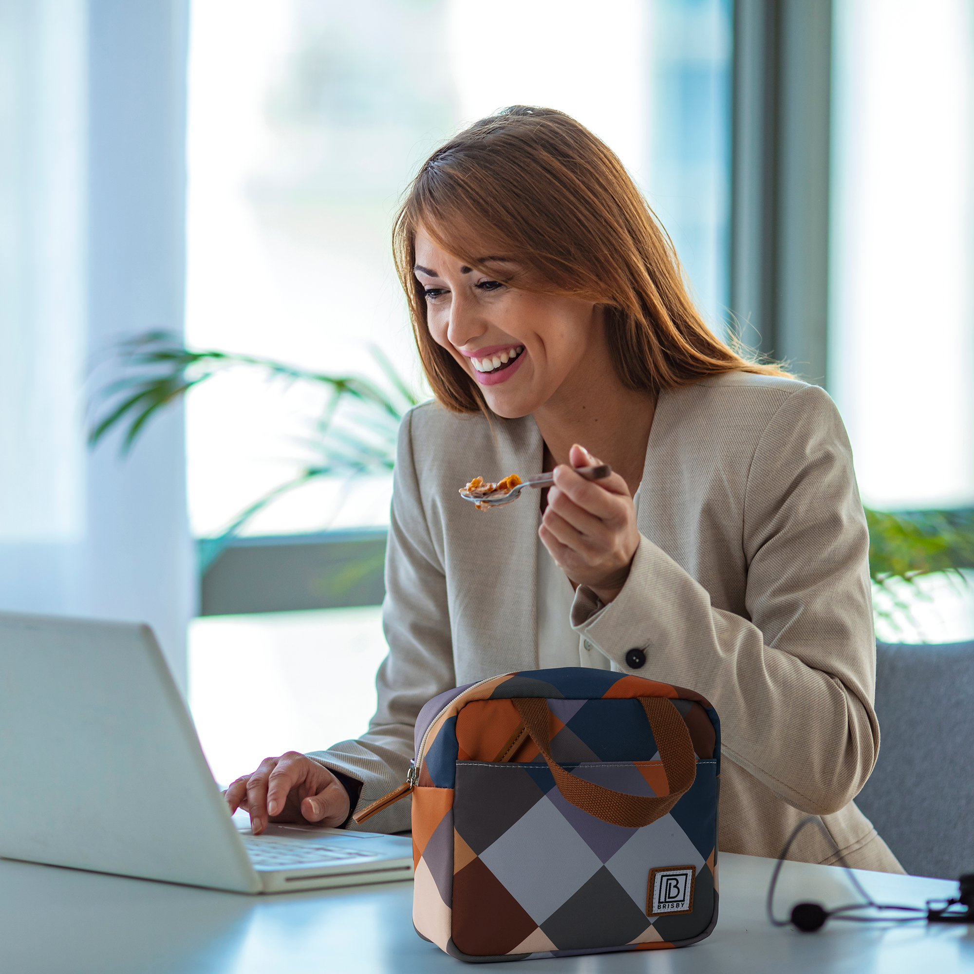 Vrouw in beige blazer eet ontbijtgranen aan een bureau met laptop en lunchtas. Lacht en werkt.