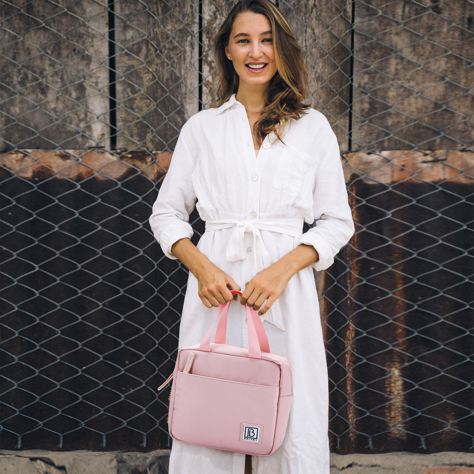 Vrouw in witte jurk lacht, houdt roze tas vast. Achtergrond: metalen hek en houten muur.