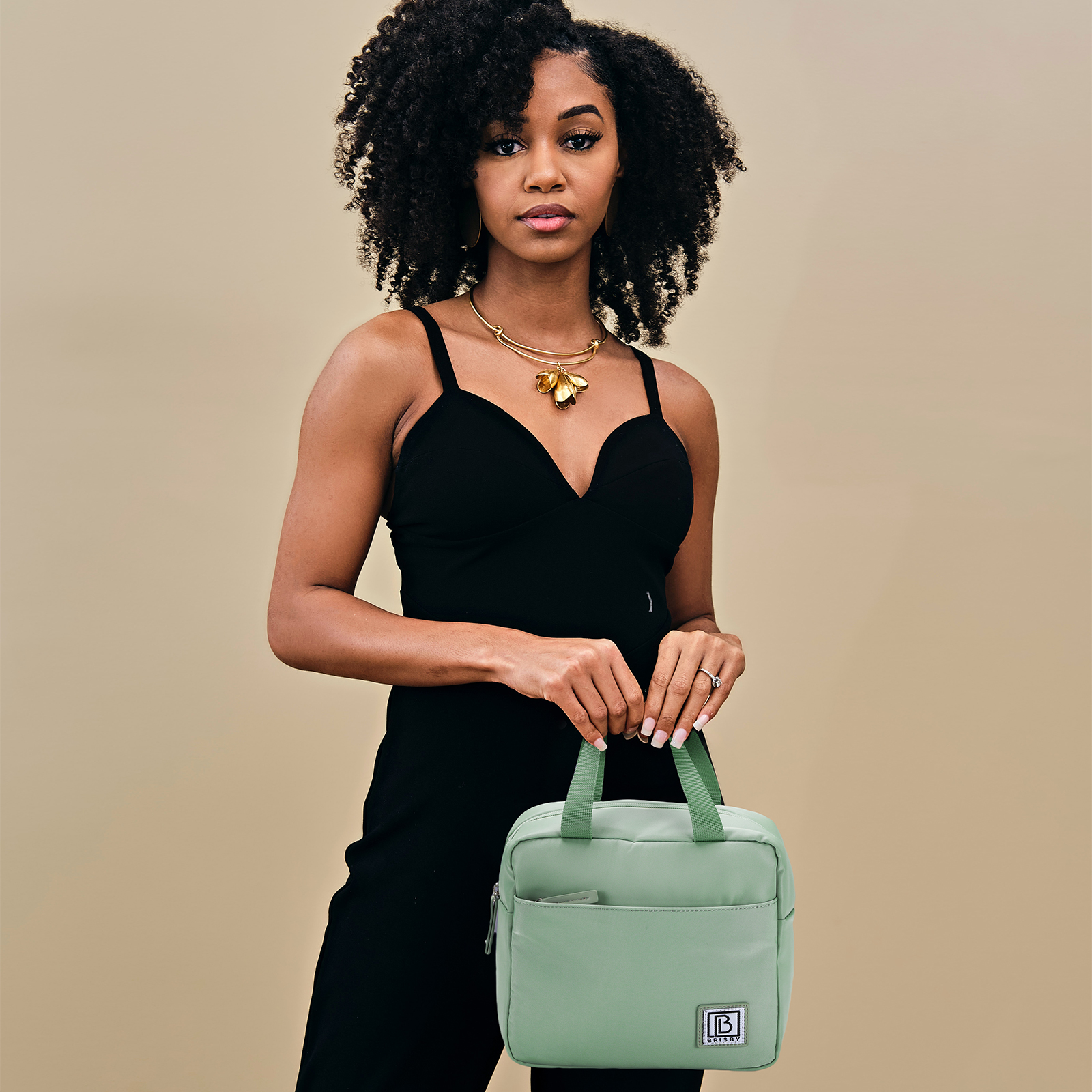 Een vrouw in een zwarte jurk houdt een groene tas vast. Ze heeft krullend haar en draagt gouden sieraden. Beige achtergrond.