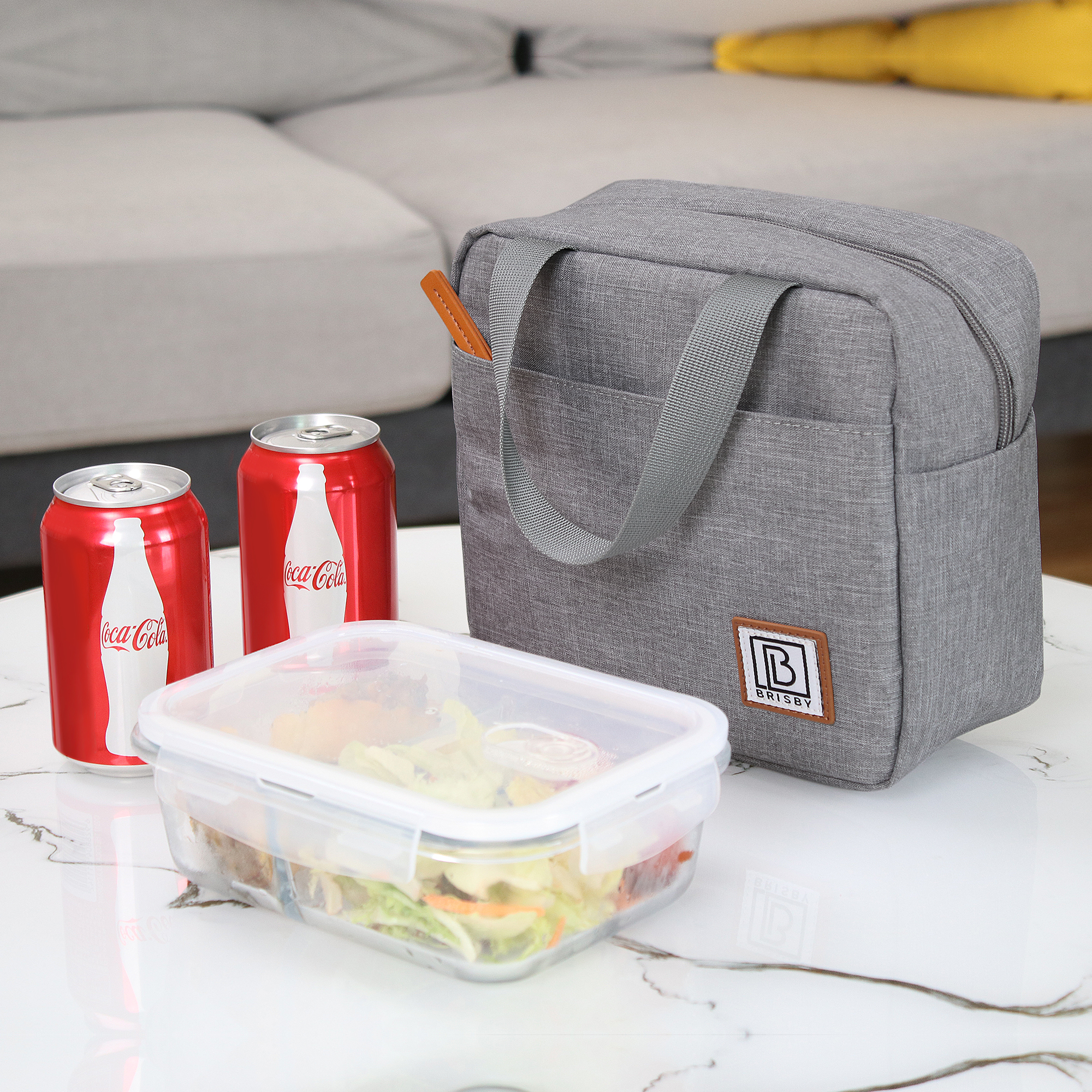 Een grijze lunchtas, twee Coca-Cola-blikjes en een bakje salade staan op een witte tafel.