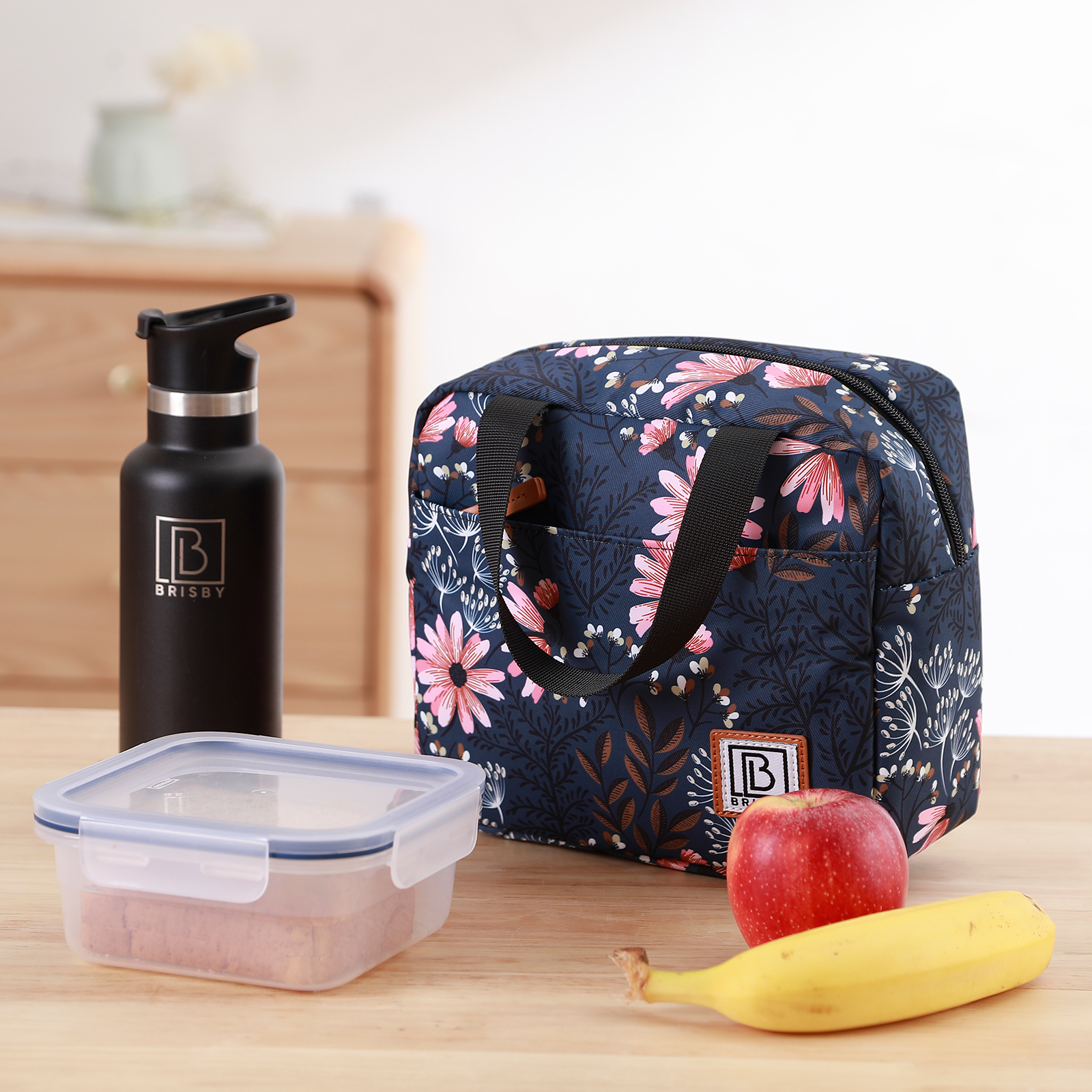 Een lunchsetting met een bloemige lunchtas, waterfles, bakje, appel en banaan op een houten tafel.