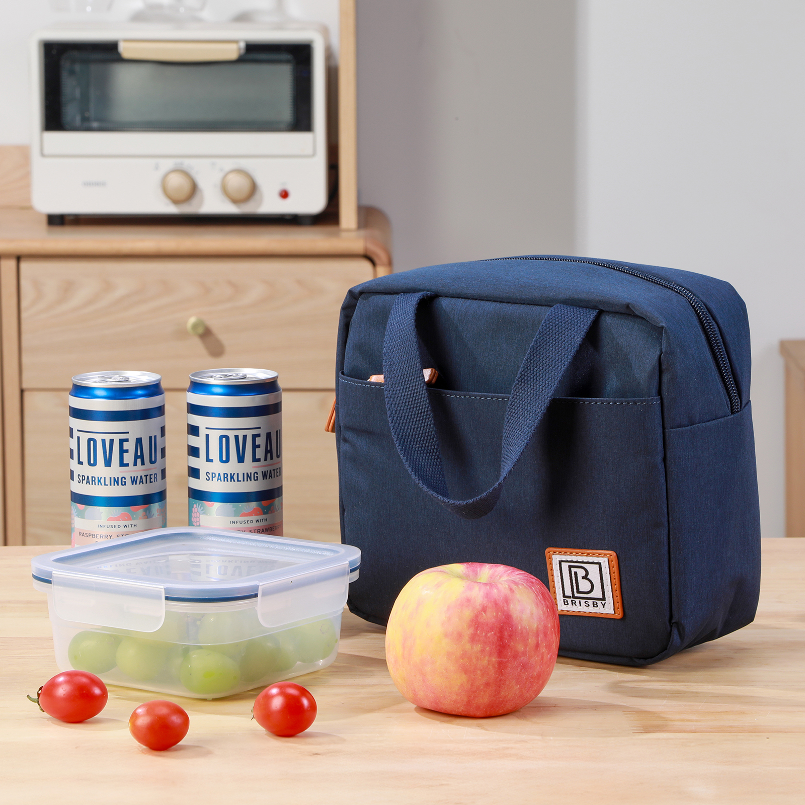 Een lunchtas, appel, tomaten, frisdrankblikjes en een bakje met druiven op een houten tafel. Een broodrooster op de achtergrond.