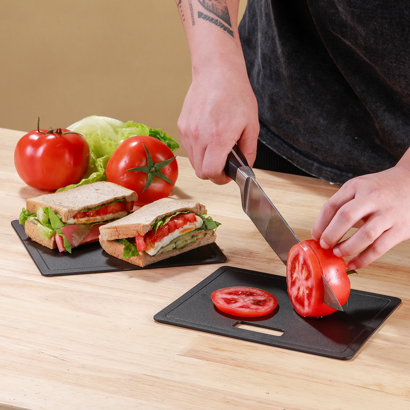 Een persoon snijdt een rode tomaat op een zwarte snijplank, sandwiches en tomaten op de achtergrond.