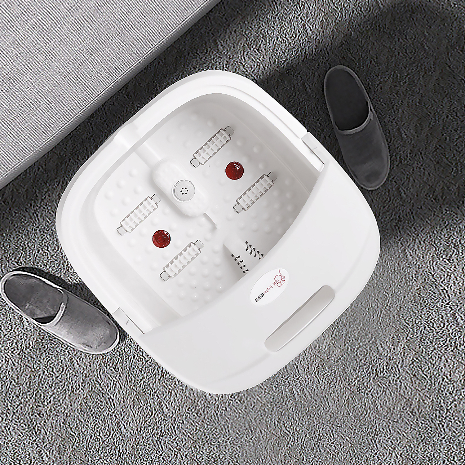 Un spa de pies blanco con detalles rojos, rodeado de dos zapatillas grises sobre una alfombra gris. Un sofá gris al fondo.