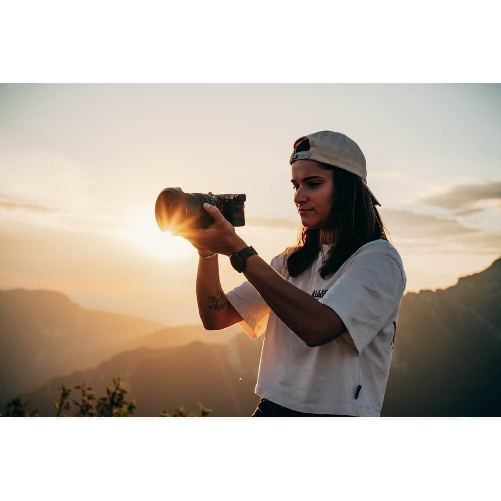 A woman holding a camera, backlit by the setting sun, capturing a mountain landscape. She wears a cap and white tee.
