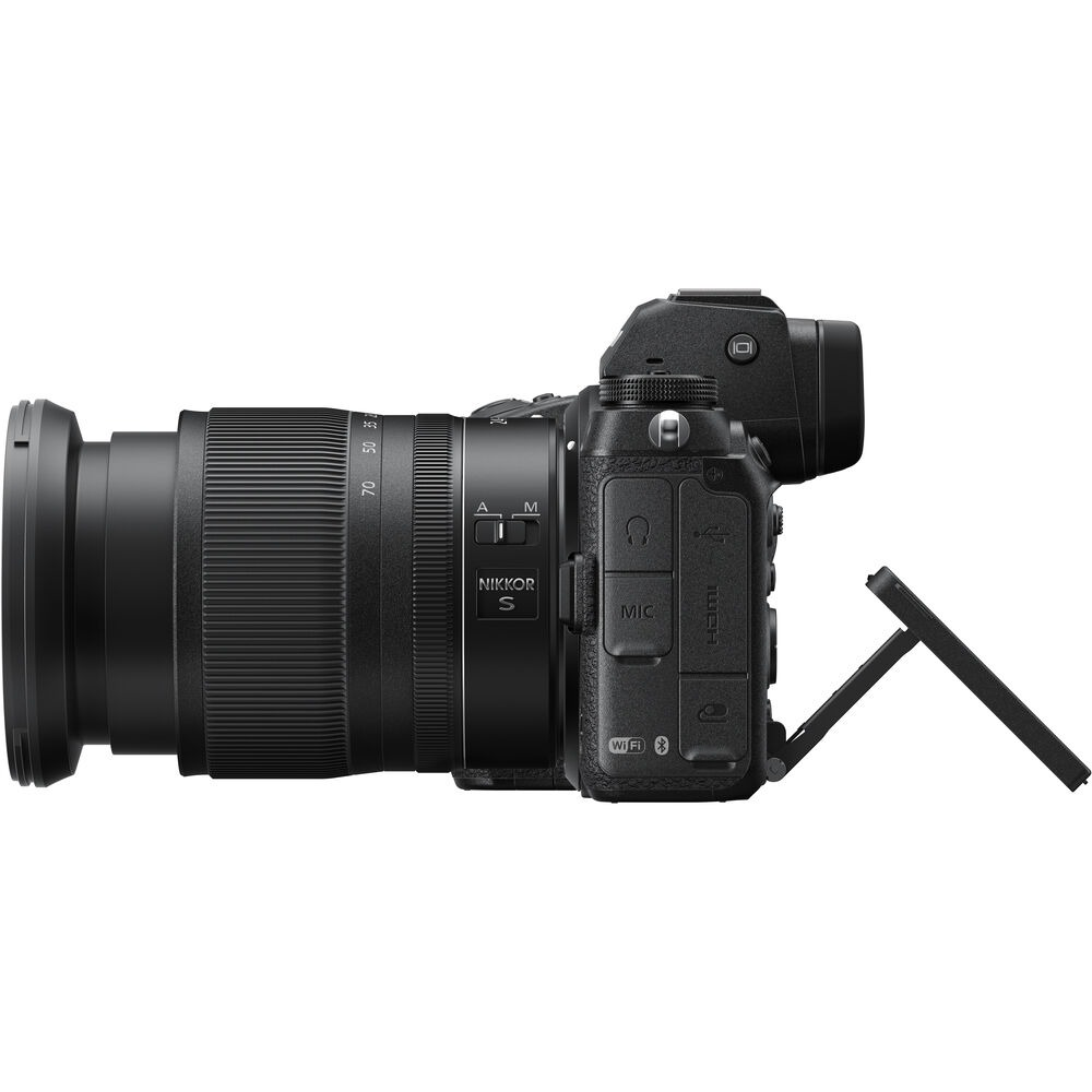 Schwarze Nikon-Kamera mit Objektiv, Seitenansicht. Das Objektiv hat Markierungen, die Kamera Anschlüsse und einen ausklappbaren Bildschirm.