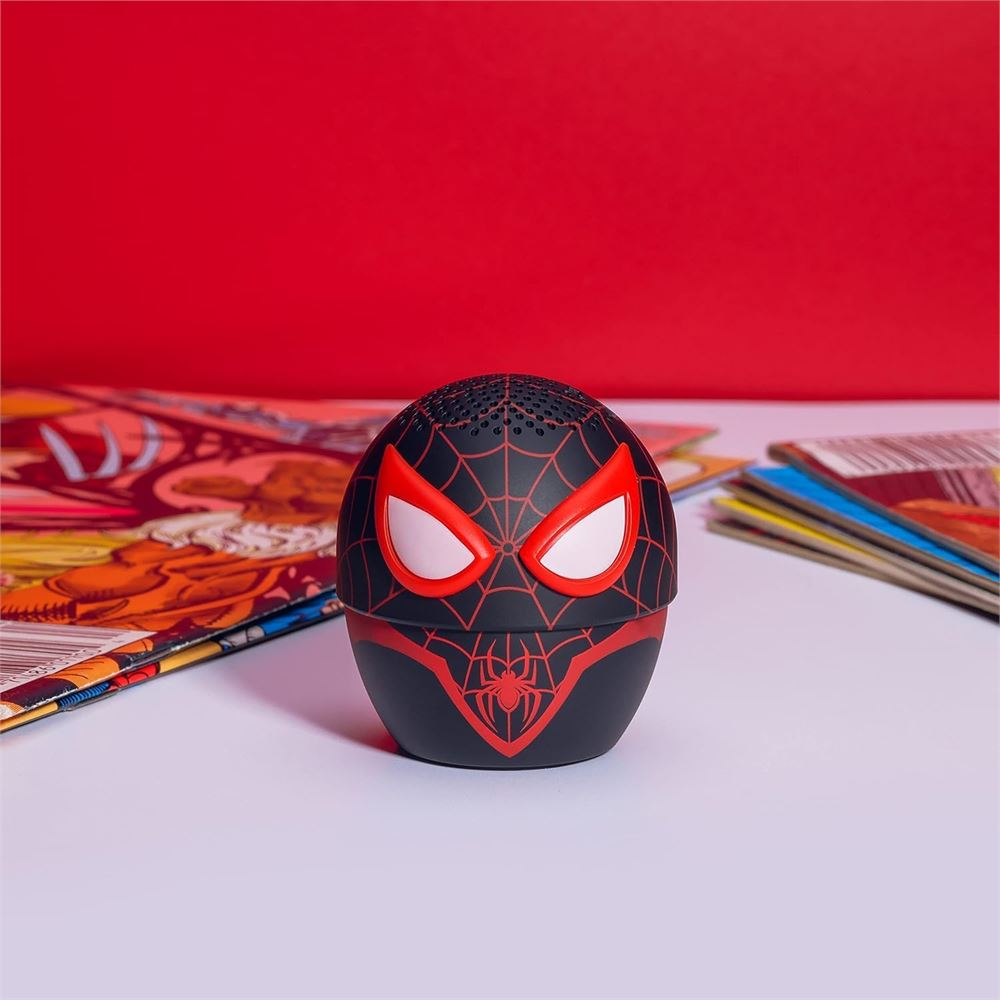 Ein Spider-Man-Lautsprecher steht auf einer weißen Oberfläche. Er hat rote Augen und ein Spinnennetz-Design. Comics sind im Hintergrund.