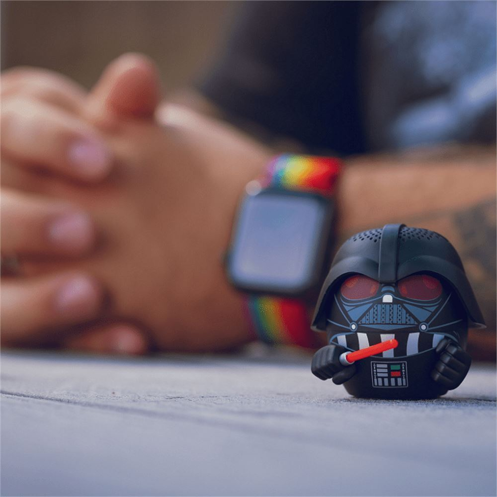 Darth Vader Figur mit rotem Lichtschwert, vor einer Person mit Regenbogenuhr. Der Hintergrund ist unscharf.