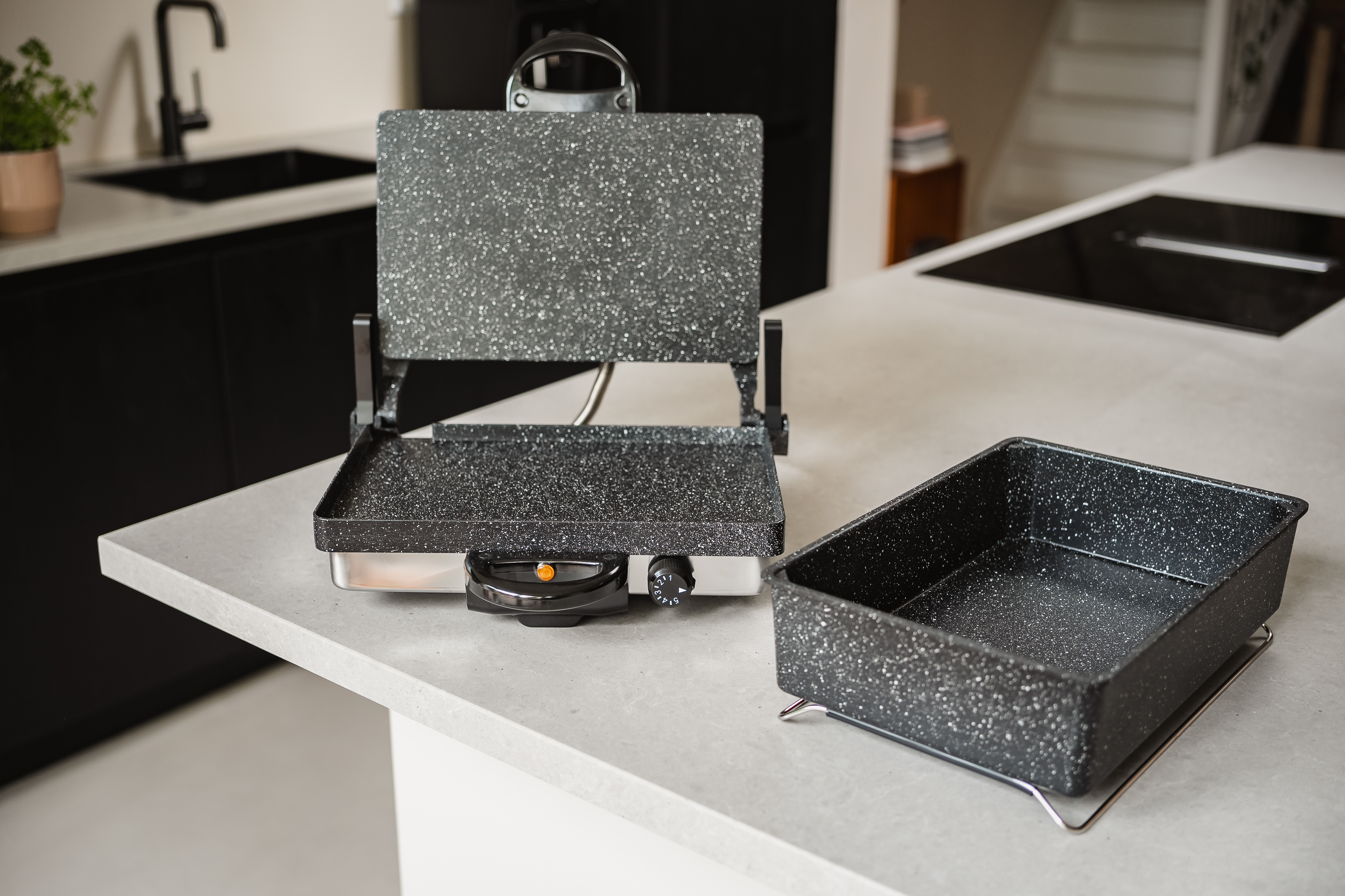 Open electric grill with a tray next to it on a kitchen counter. Black sink and a stove visible.