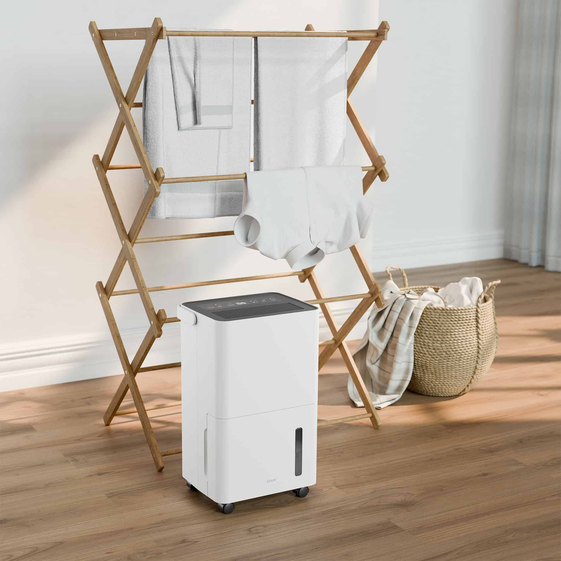 Een witte luchtontvochtiger op wielen, een houten droogrek met handdoeken en kleding, een rieten mand met een deken.