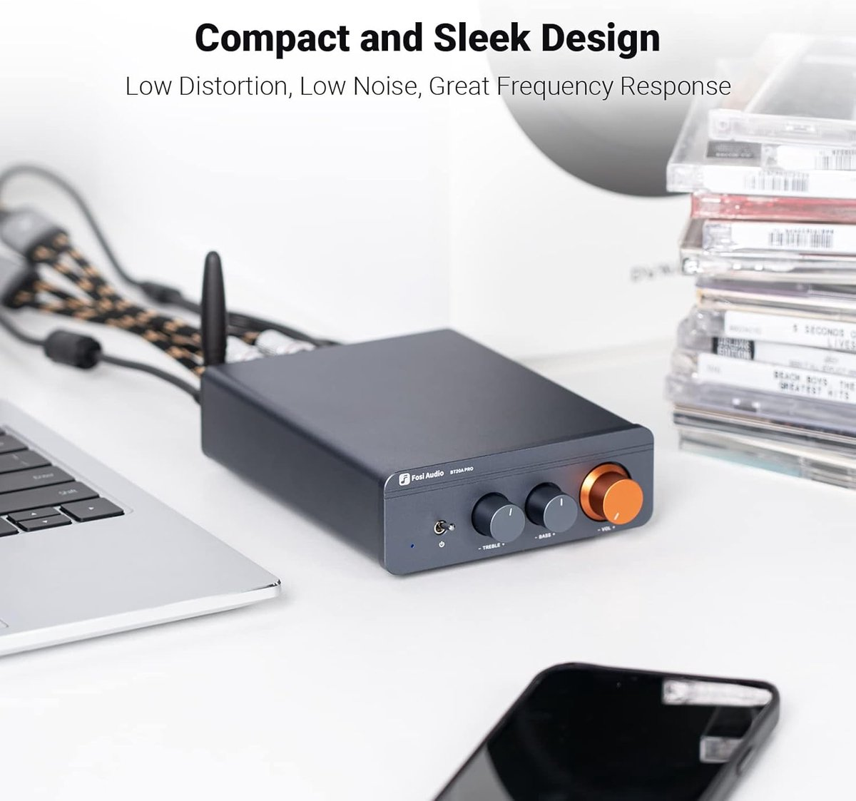 Un amplificateur audio gris foncé avec des boutons est posé sur une table blanche à côté d'un ordinateur portable, de câbles et de CD. Un téléphone est au premier plan.
