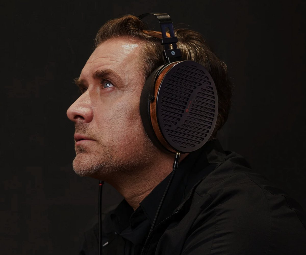 Man wearing headphones looking upwards. Dark background. Headphones are black with wooden accents. Man is fair-skinned with short hair.