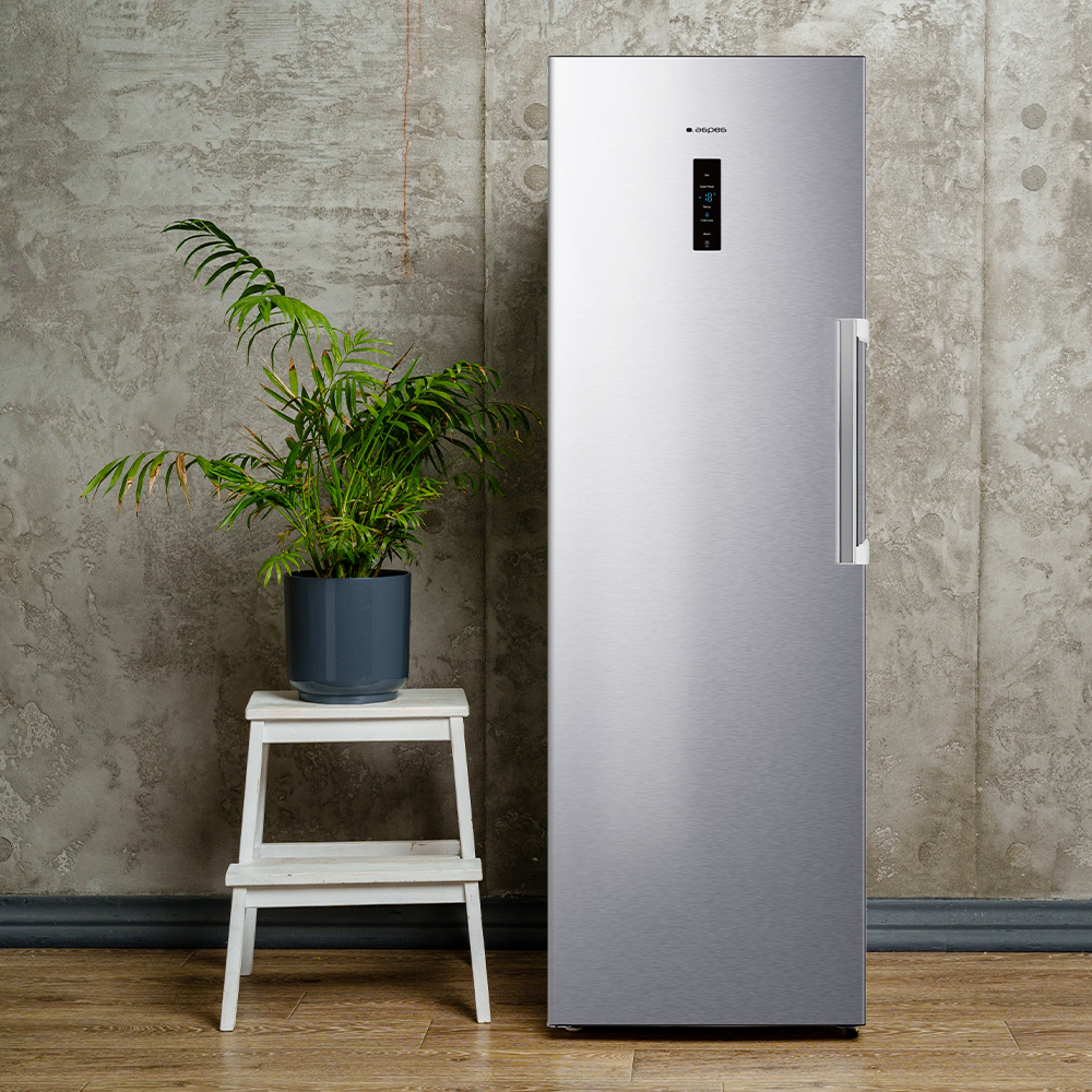 Un refrigerador de acero inoxidable junto a una planta en un taburete blanco. El fondo es una pared gris texturizada y suelo de madera.