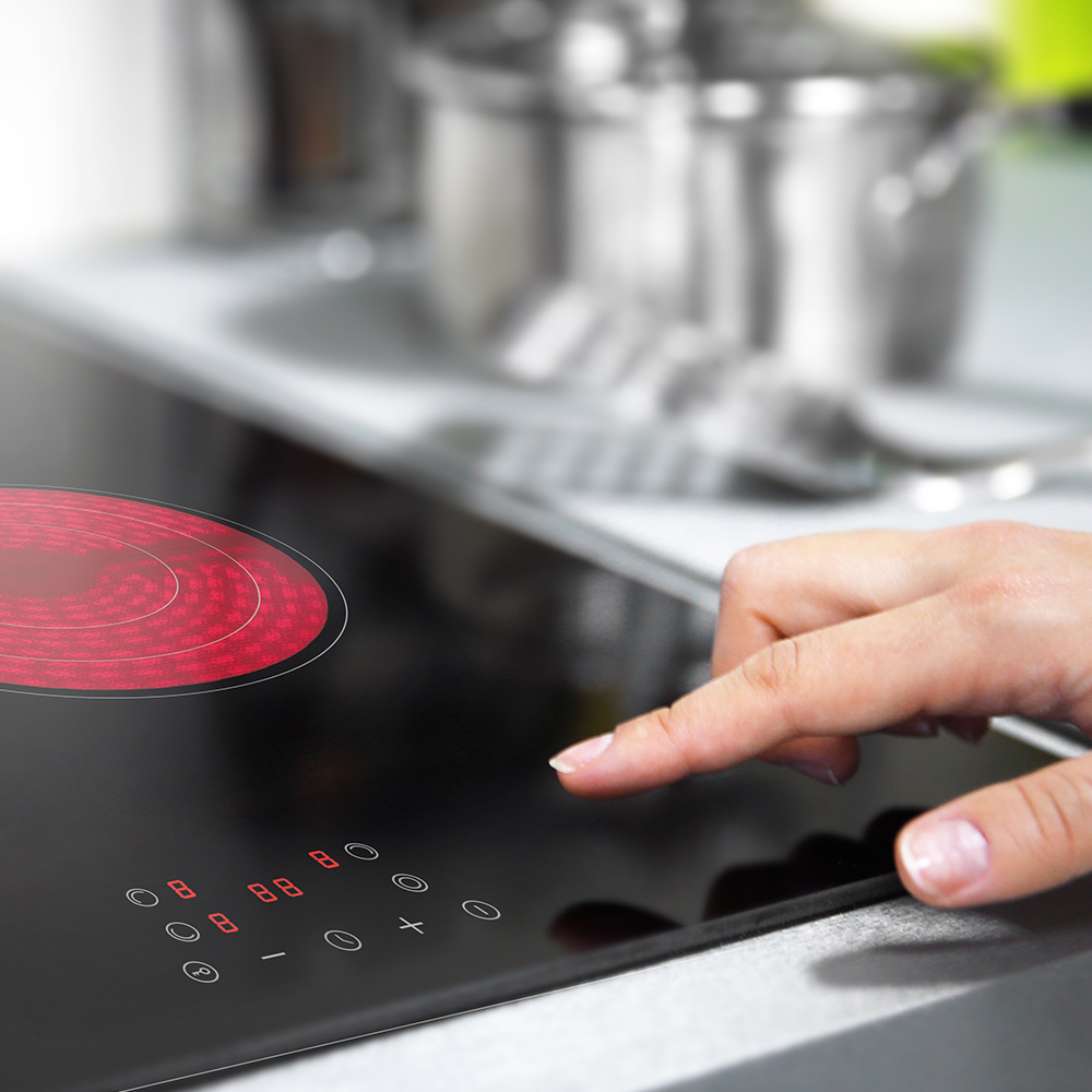 Ręka dotyka czarnej płyty grzejnej z czerwonym elementem grzewczym. Wyświetlacz cyfrowy pokazuje liczby. Tło: przybory kuchenne i stal nierdzewna.