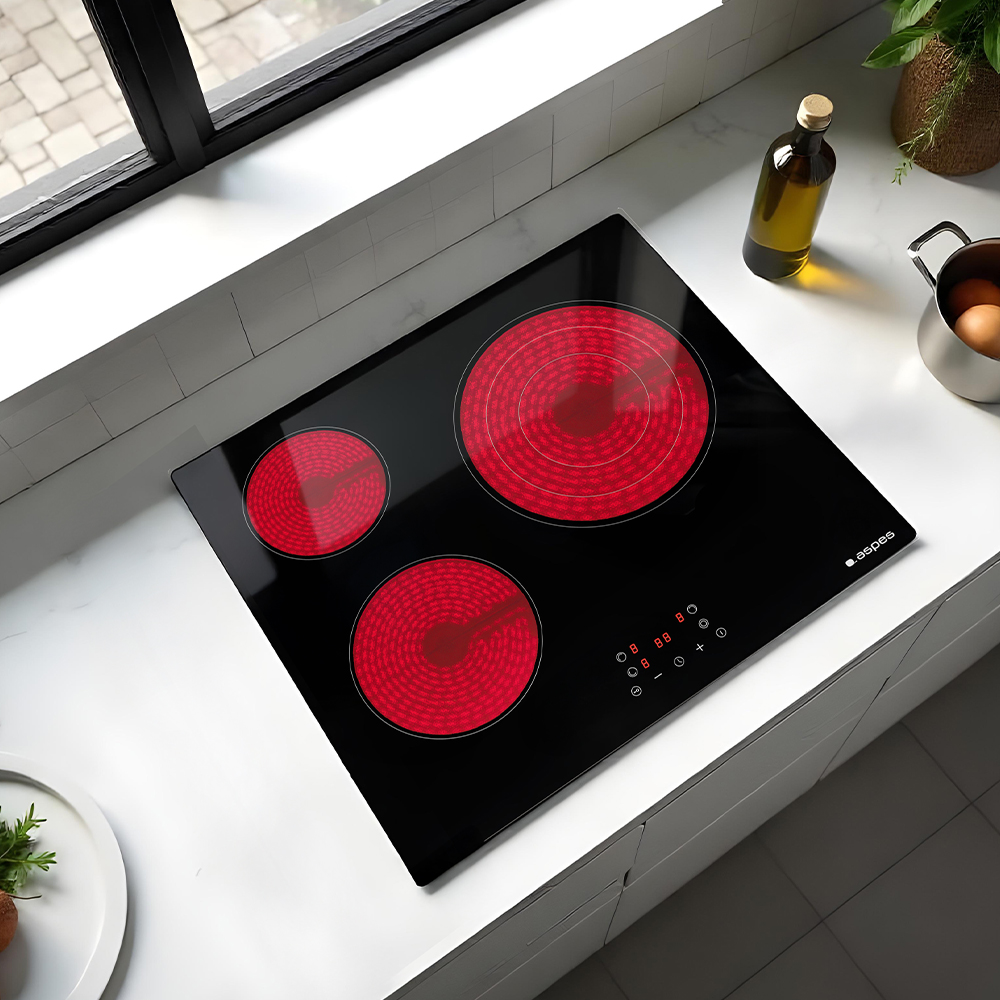 Czarna płyta grzewcza z trzema czerwonymi palnikami. Butelka oliwy z oliwek, garnek z jajkami i roślina na białym blacie.