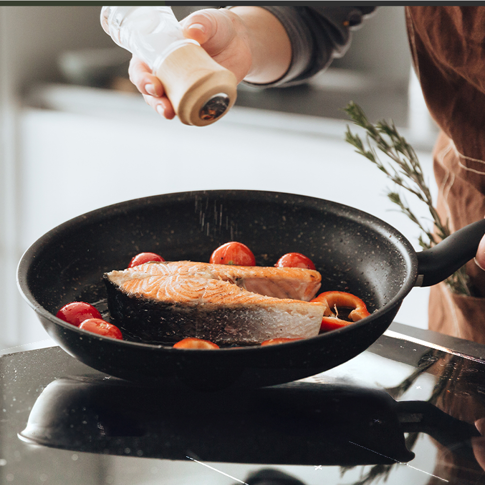 Osoba doprawia łososia solą z młynka na patelni z pomidorami i papryką. Widoczny jest brązowy fartuch.