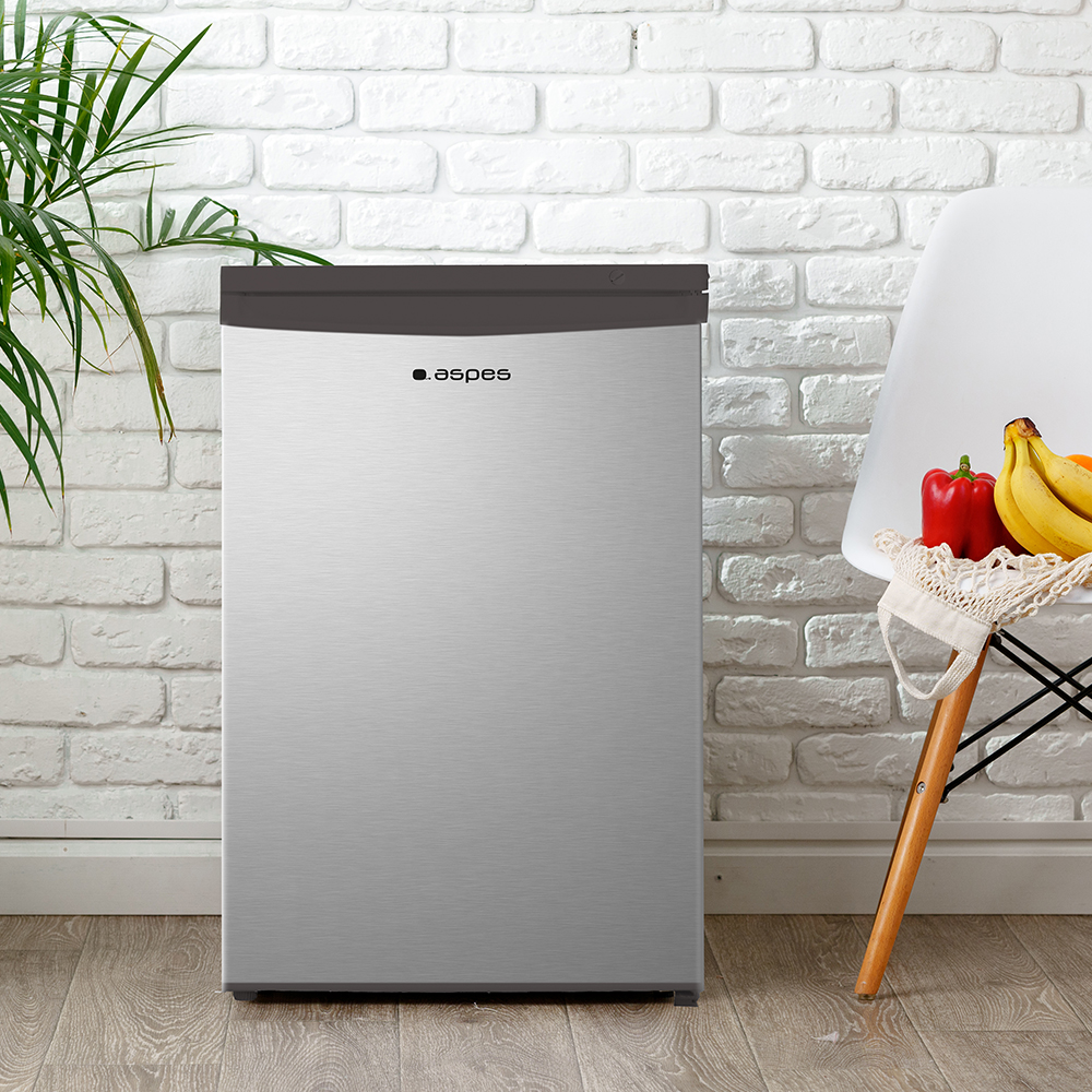 Un mini-refrigerador plateado contra una pared de ladrillos blancos. Una silla con frutas. Una planta a la izquierda.