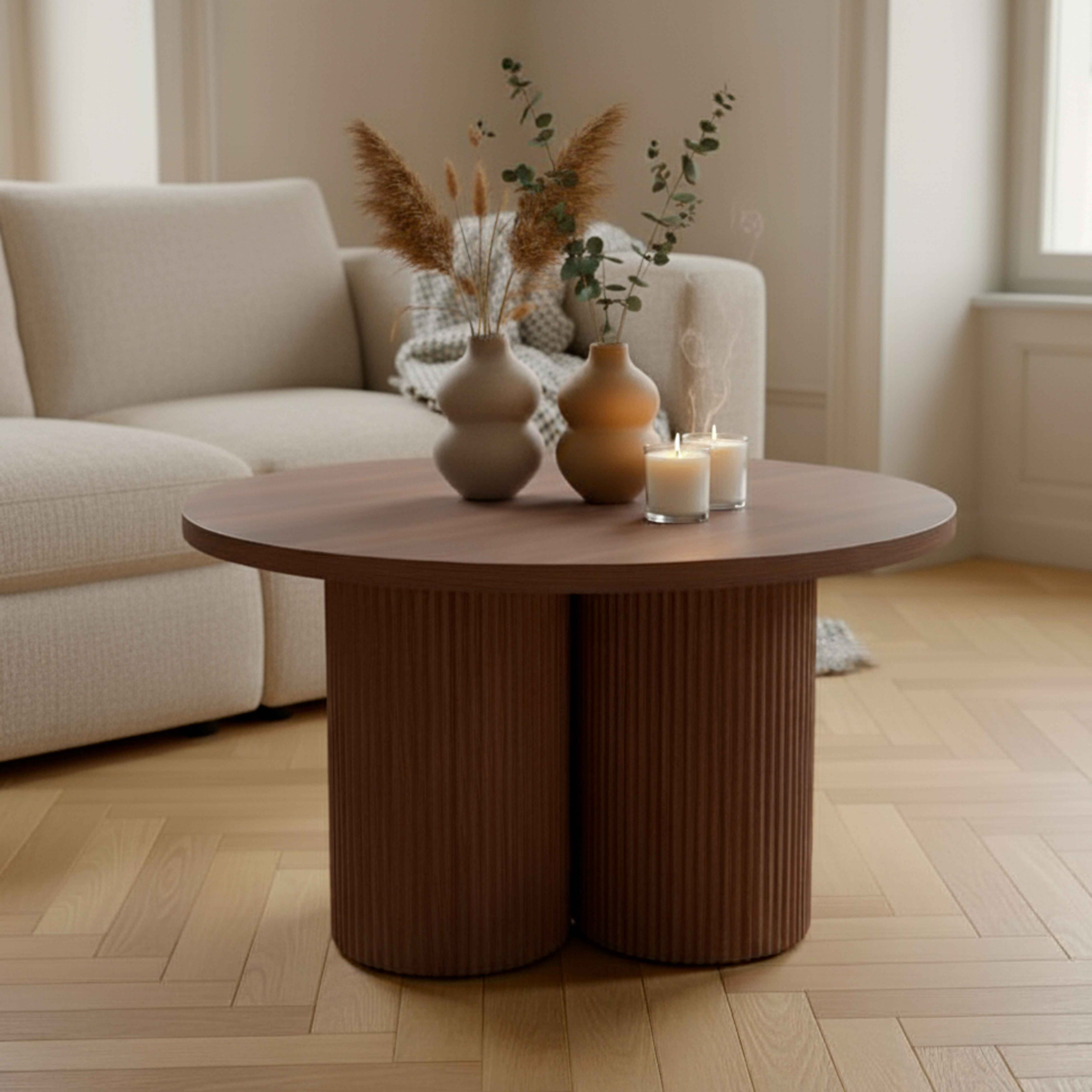 Runder brauner Tisch mit Vasen und Kerzen. Beiges Sofa im Hintergrund. Holzfußboden mit Fischgrätmuster.
