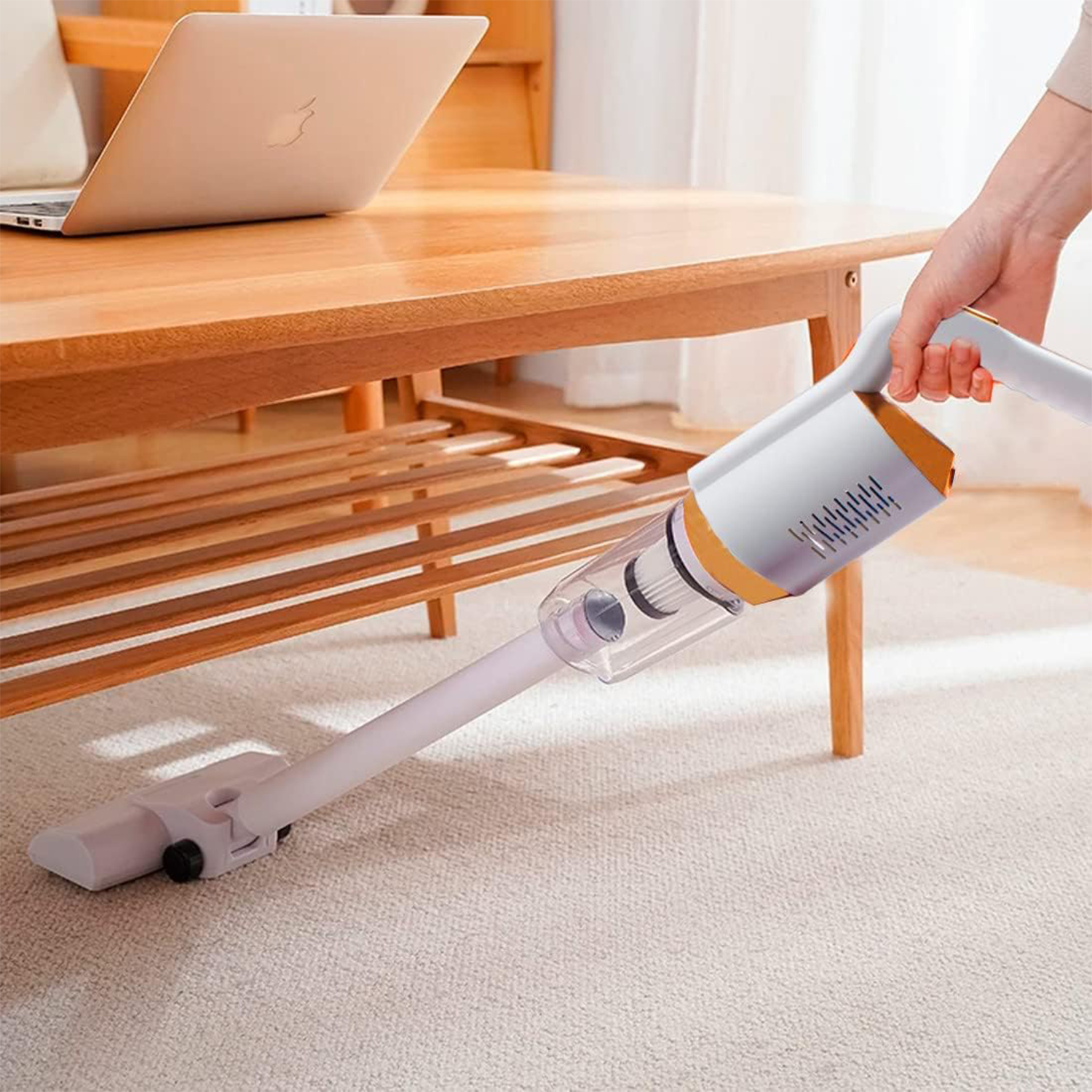 Una persona usa una aspiradora de mano blanca y naranja en una alfombra. Una computadora portátil está sobre una mesa de madera.