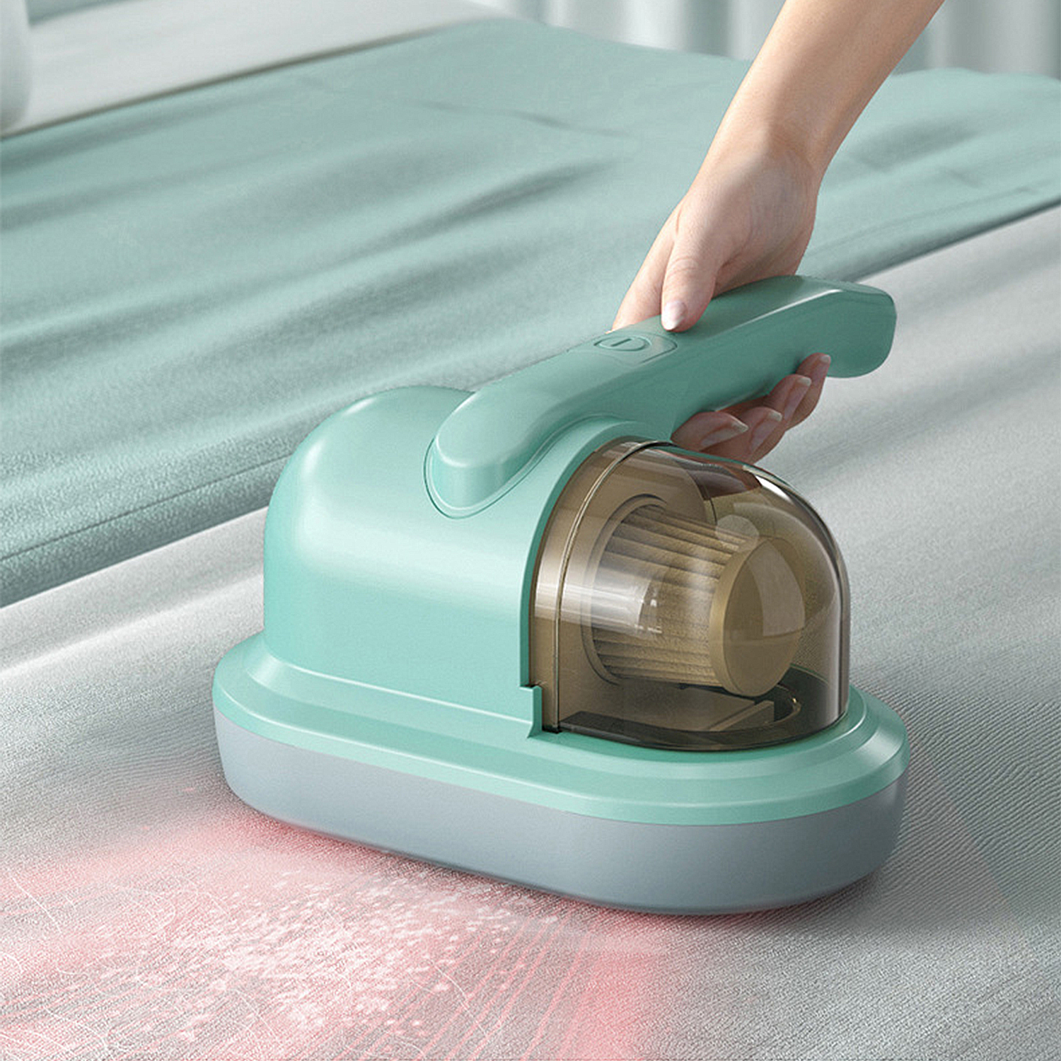 Una mano sostiene una aspiradora de mano verde azulado sobre una cama. La aspiradora está limpiando la cama, luz roja visible.