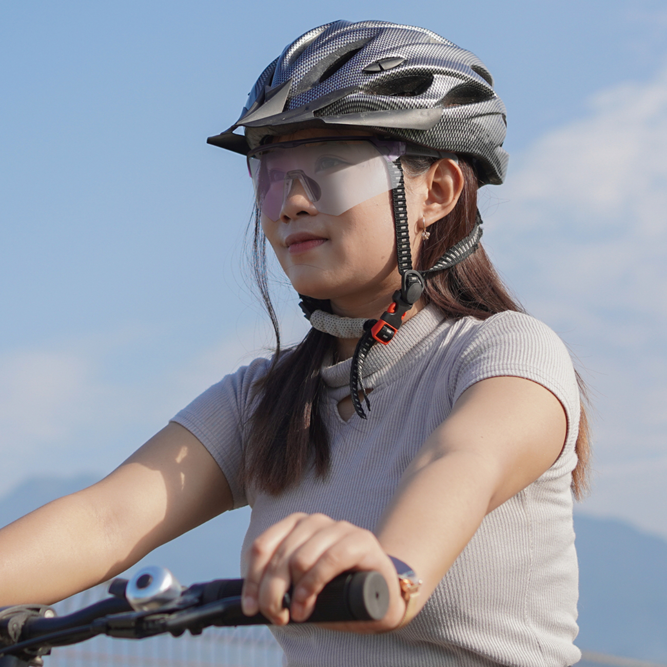 Eine Frau mit Helm und Brille, die einen Lenker hält. Blauer Himmel im Hintergrund. Berge sind in der Ferne sichtbar.