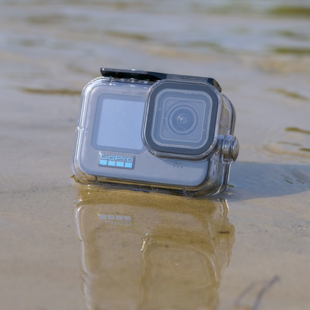 Een GoPro-camera in een doorzichtige waterdichte behuizing ligt op nat zand. De camera is grijs met het GoPro-logo. Water omringt het.