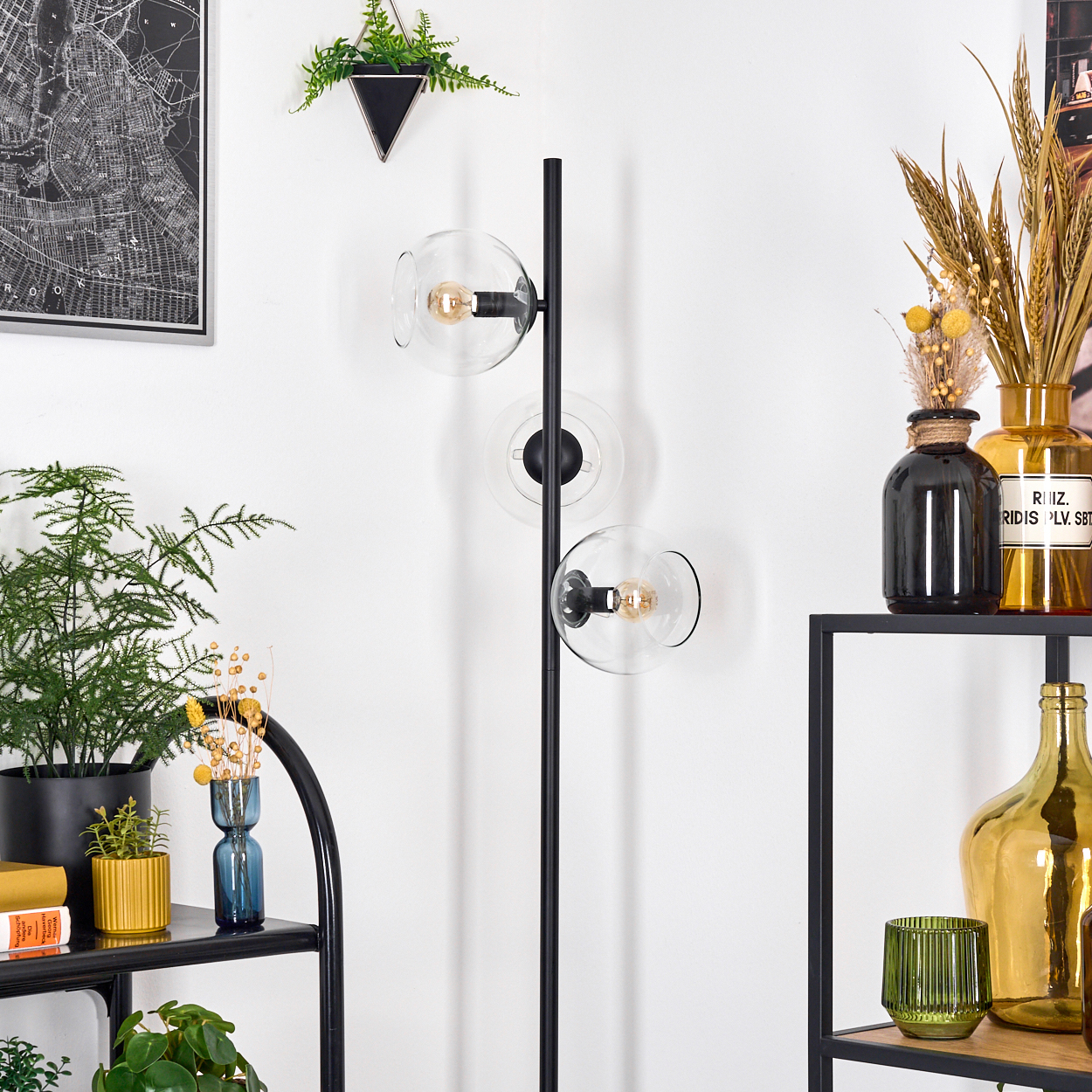 Ein moderner Wohnraum mit einer schwarzen Stehlampe mit Glaskugeln. Regale zeigen Pflanzen und dekorative Vasen vor einer weißen Wand.